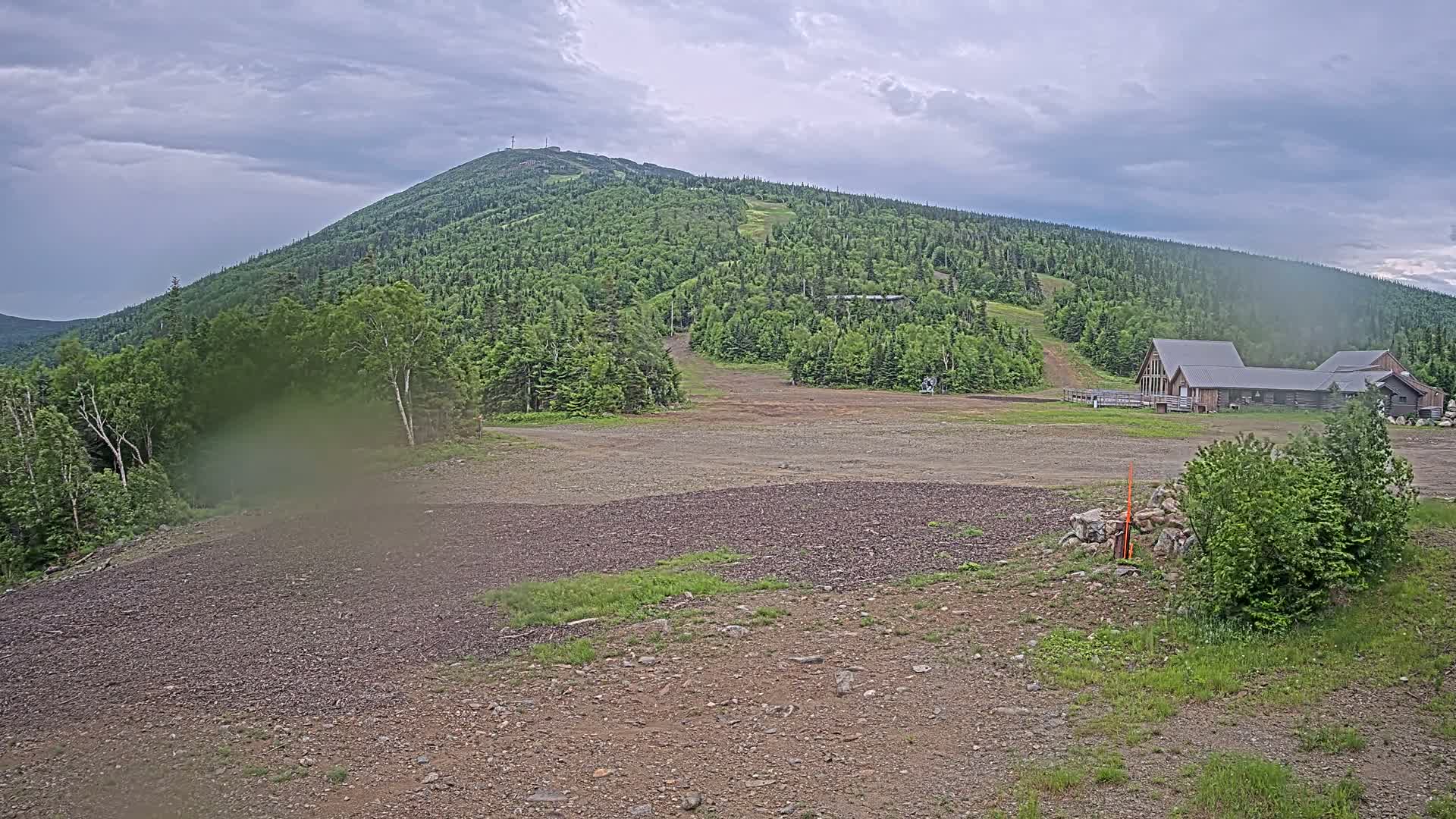 A large, forested mountain is visible under an overcast sky, with a building complex at its base and a gravel area in the foreground.