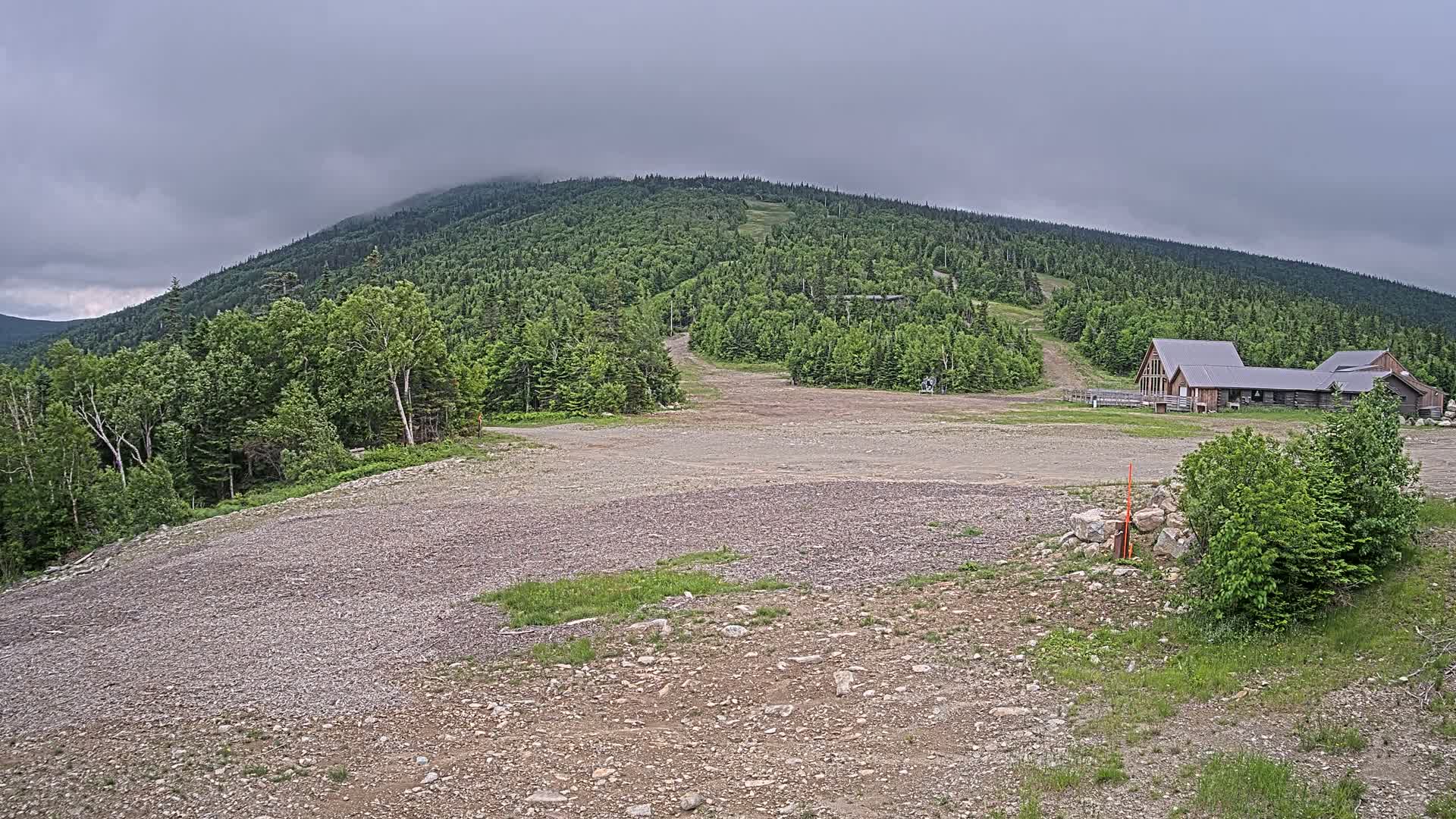 A rocky, empty area in front of a large, wood-framed building sits at the base of a verdant, fog-covered mountain under an overcast sky.