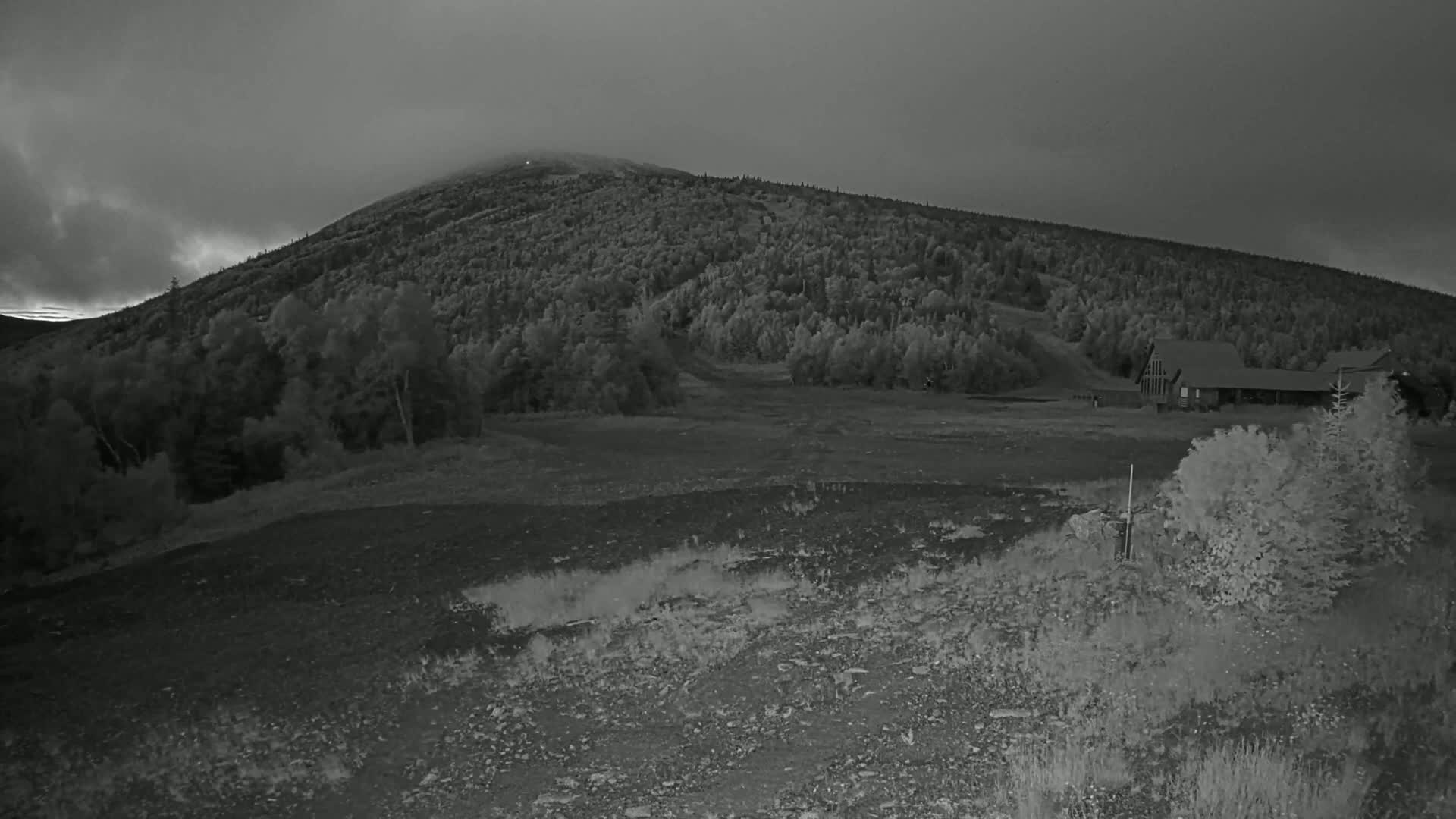 A large, forested mountain is visible under a cloudy, overcast sky, with a long building situated in a clearing at its base.