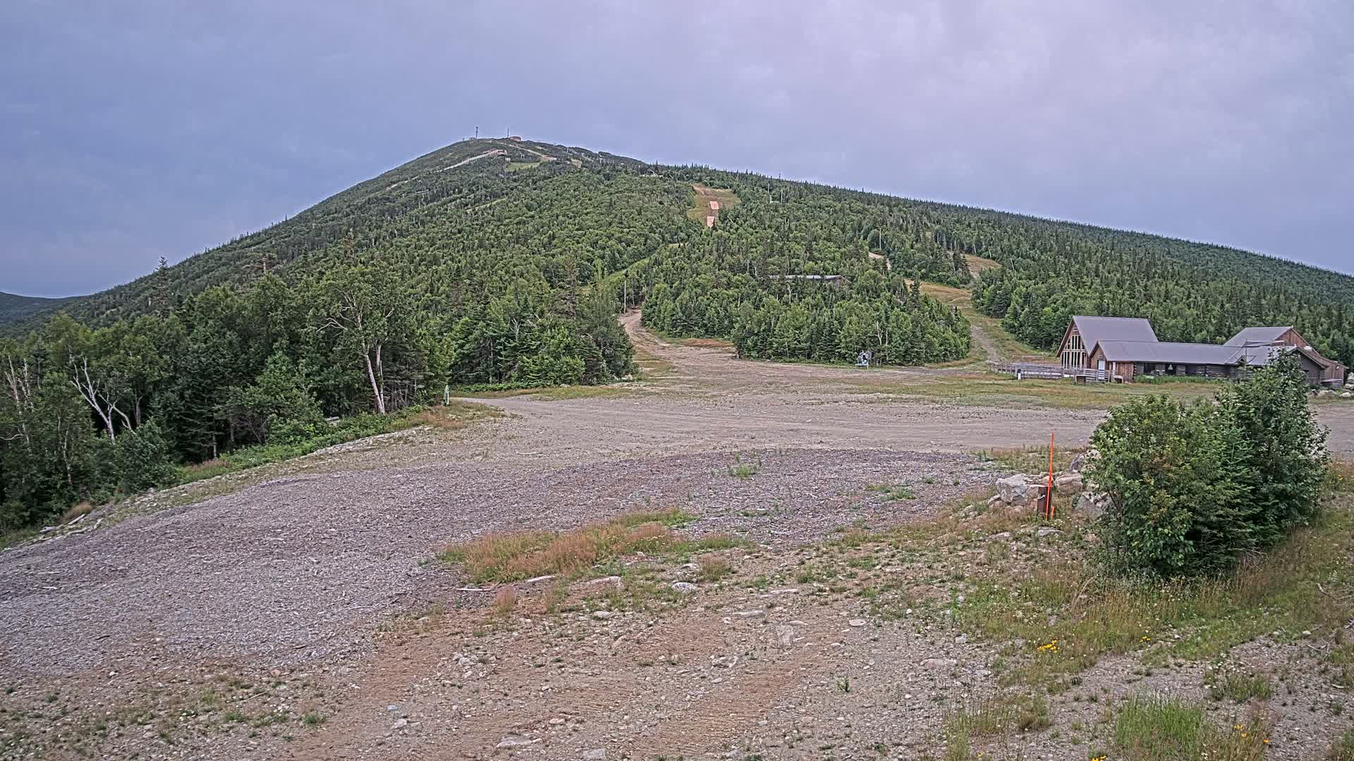 A gravel area sits in the foreground, leading to a ski resort nestled at the base of a heavily wooded mountain under a cloudy sky.