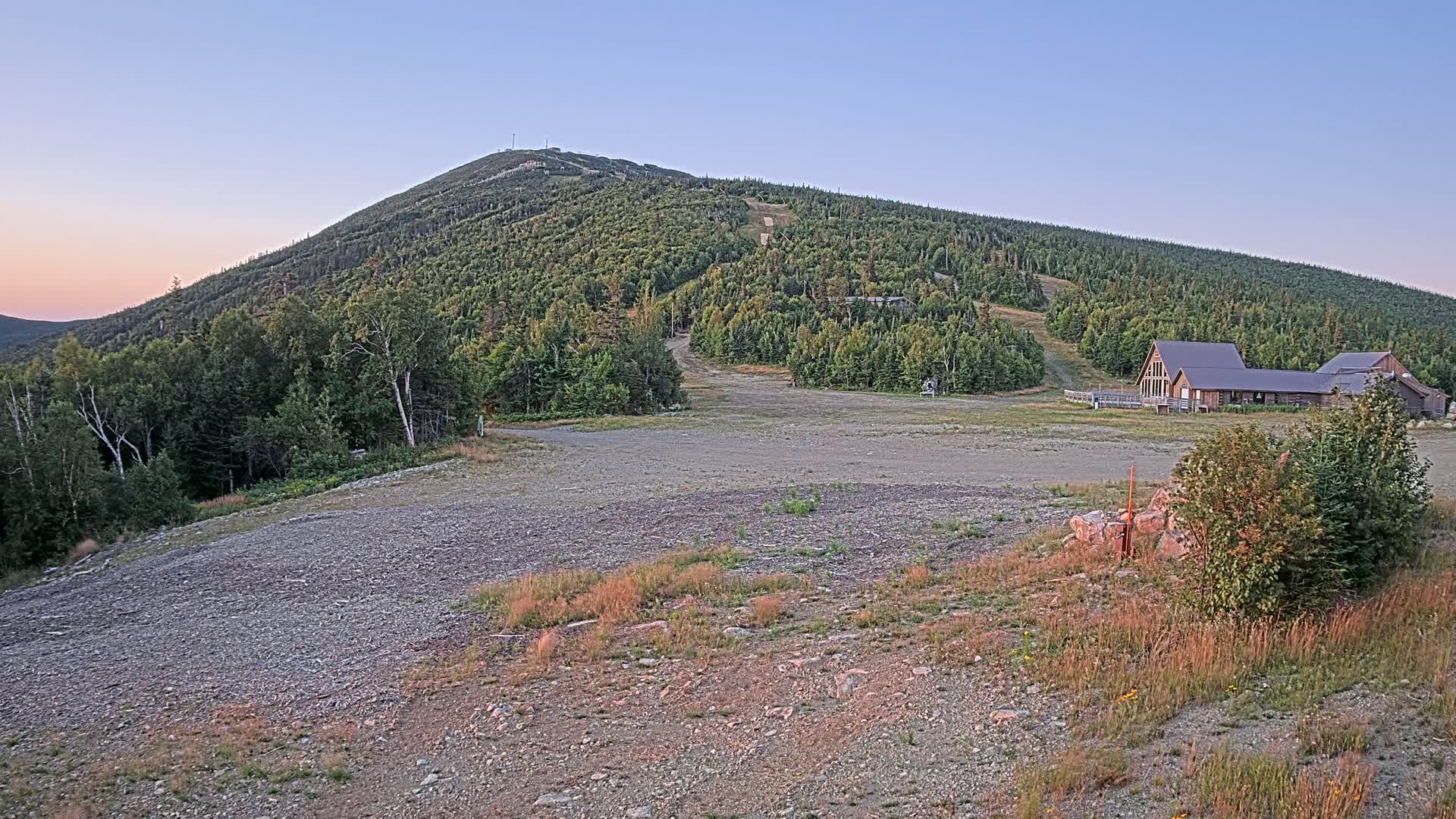 A gravel area sits in the foreground, leading to a ski resort nestled at the base of a heavily wooded mountain under a cloudy sky.