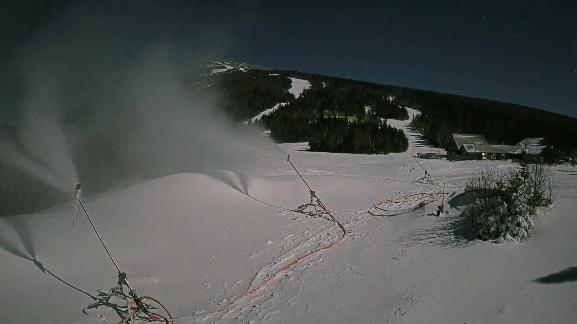 It is a clear, cold night at a snow-covered ski resort where snow guns are actively producing snow, with distant evergreen forests and lodge buildings visible under a starry, moonlit sky.