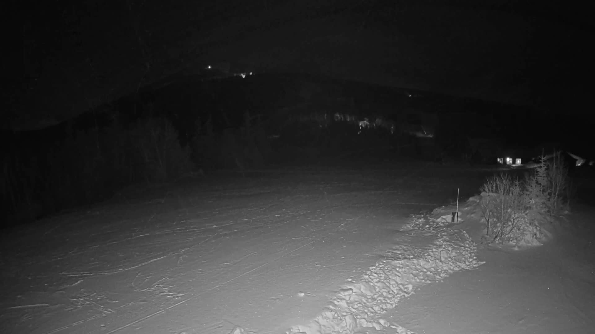 It is a clear, cold night at a snow-covered ski resort where snow guns are actively producing snow, with distant evergreen forests and lodge buildings visible under a starry, moonlit sky.