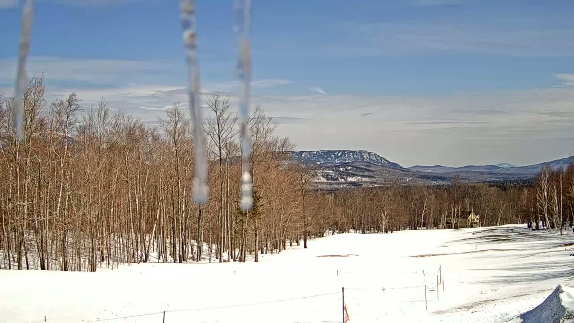 A snow-covered landscape with bare trees and a distant mountain range under a partly cloudy, sunny sky is viewed through melting icicles.