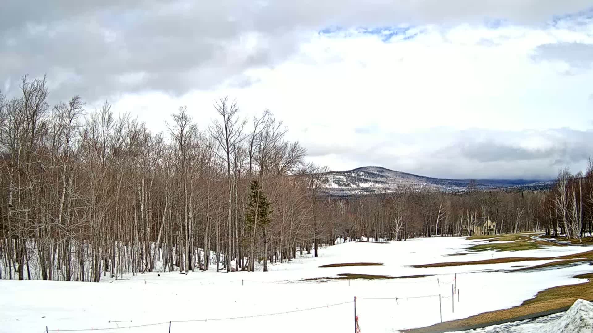 A partly cloudy day shows a snow-covered golf course with bare trees in the foreground and a snow-capped mountain range in the distance.