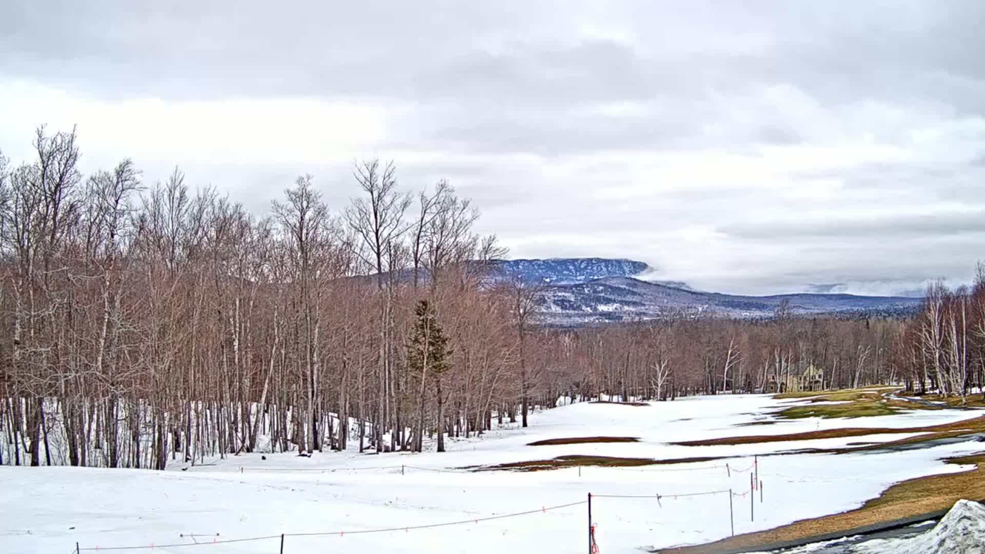 A snow-covered golf course is nestled amongst bare, winter trees, with a mountain range partially obscured by clouds in the background under a cloudy sky.