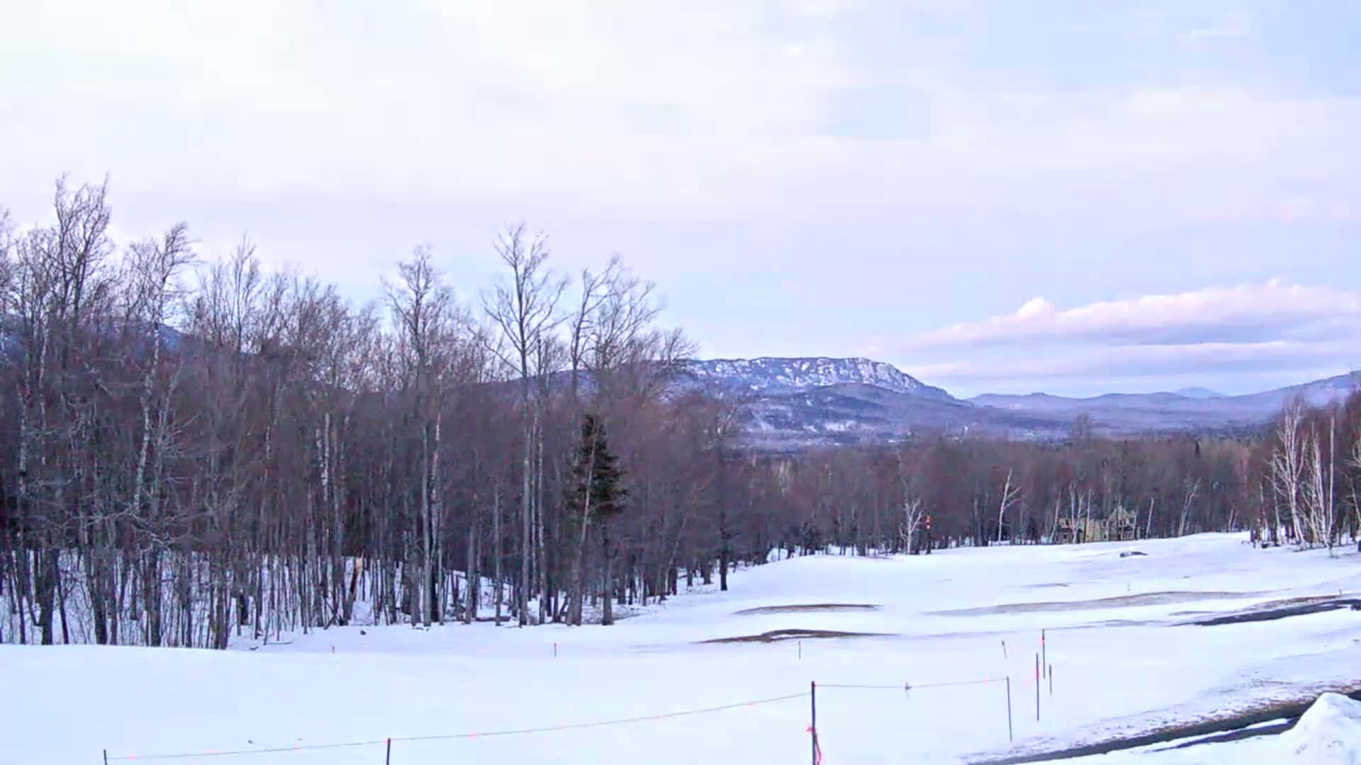 A snow-covered golf course stretches out before a line of bare trees, with distant mountains under a pale sky.
