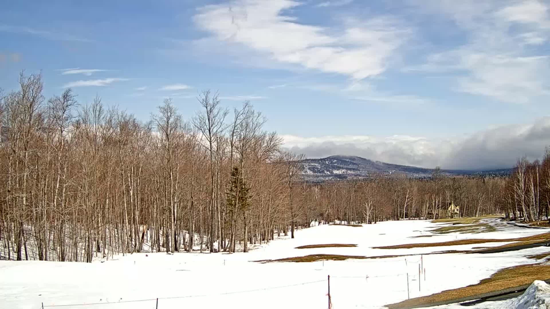 A partly cloudy day reveals a snow-covered golf course bordered by bare trees, with distant mountains partially obscured by clouds in the background.