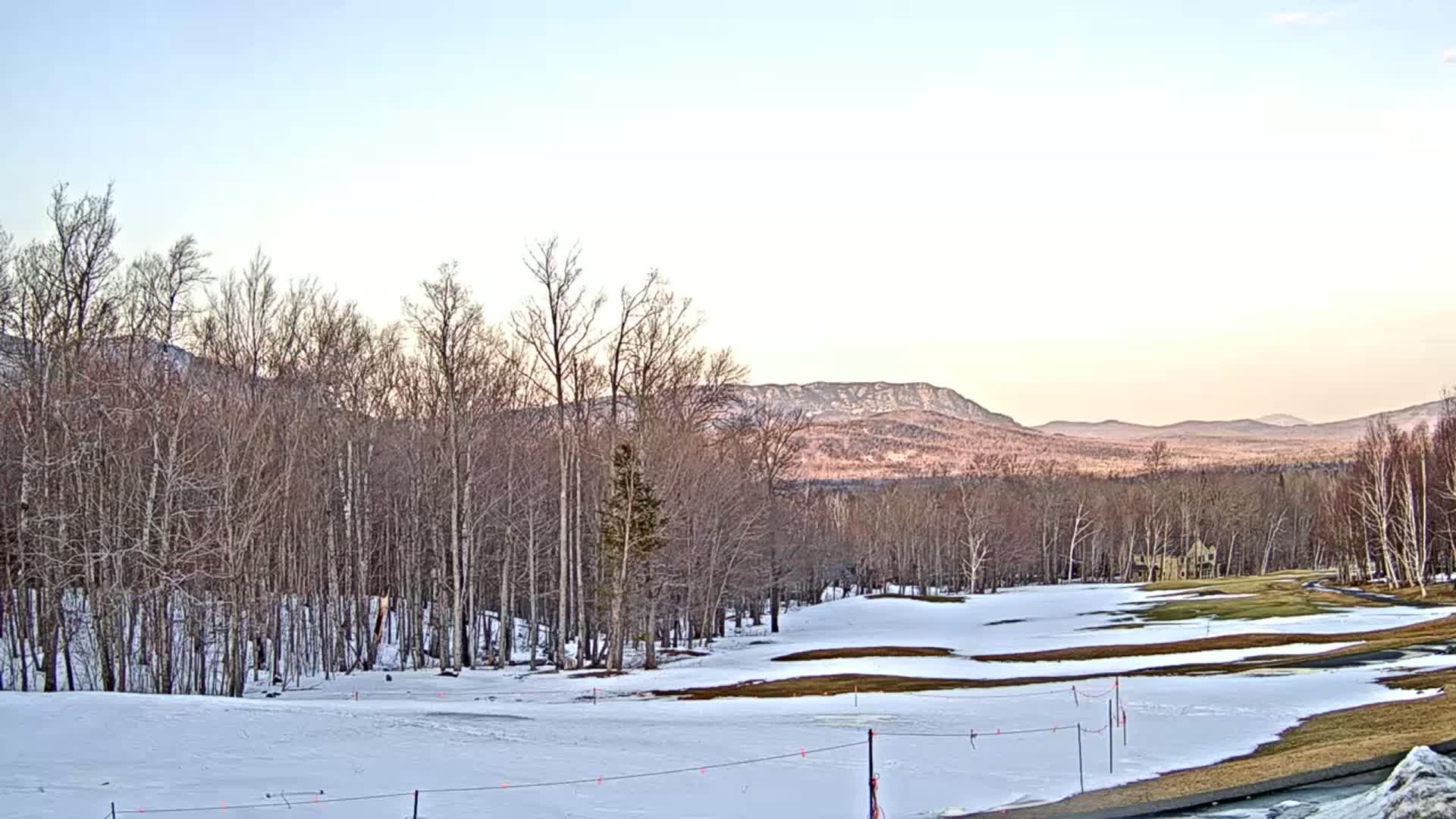 A snow-covered golf course with bare trees and distant mountains under a clear, pale sky.
