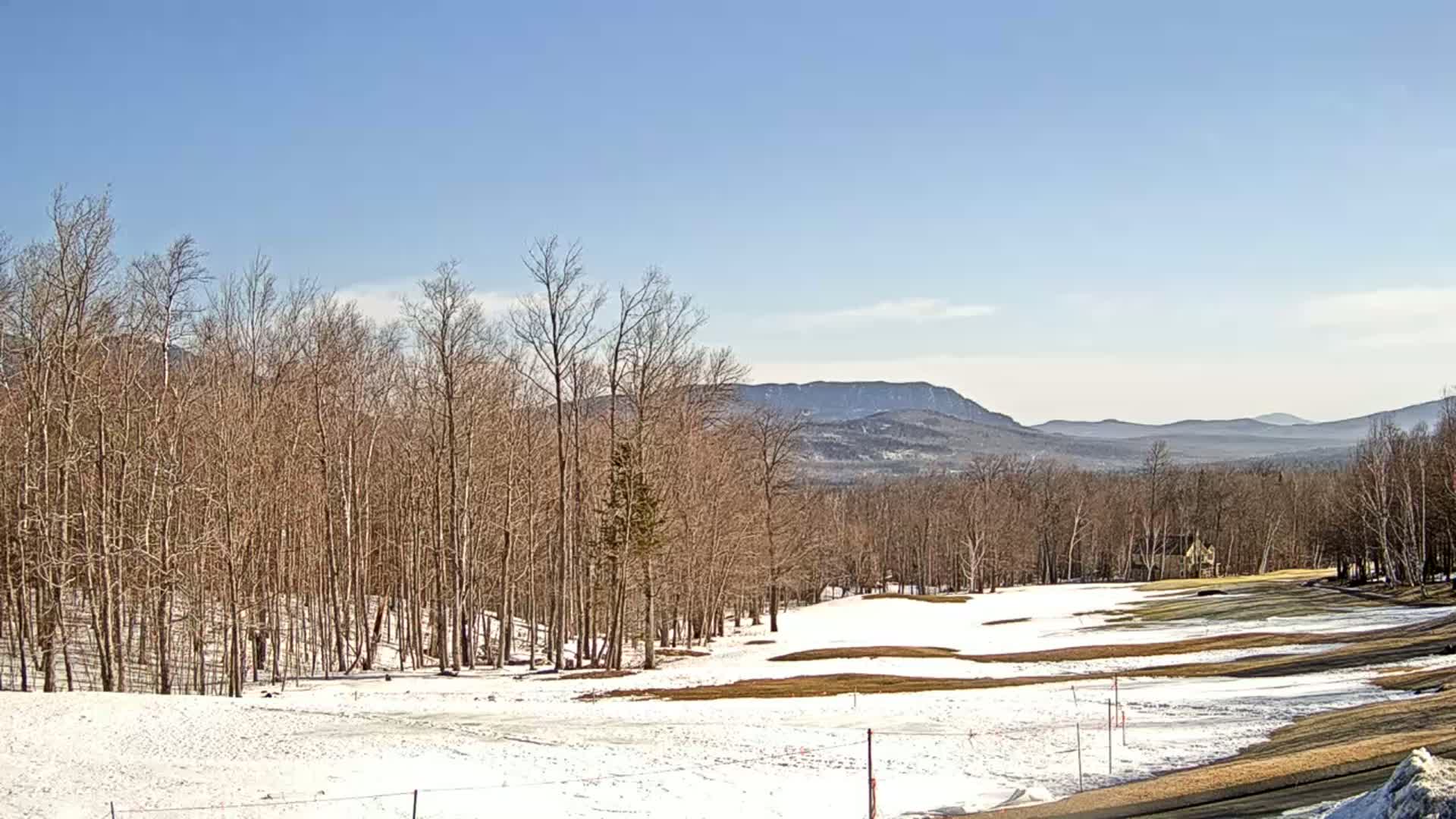 A partly snow-covered golf course with bare trees in the foreground, mountains in the background, under a clear blue sky.