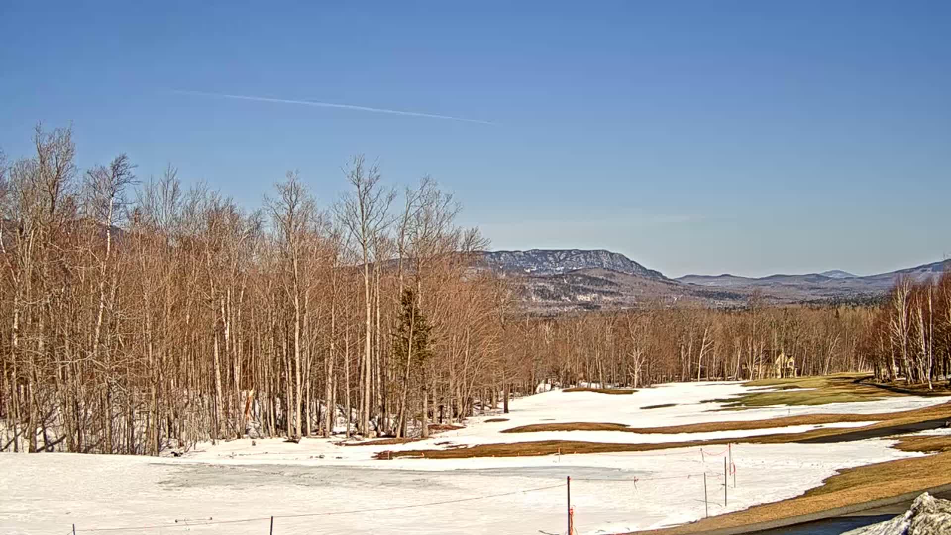 A partly snow-covered golf course with bare trees in the foreground extends to distant, snow-capped mountains under a clear blue sky.