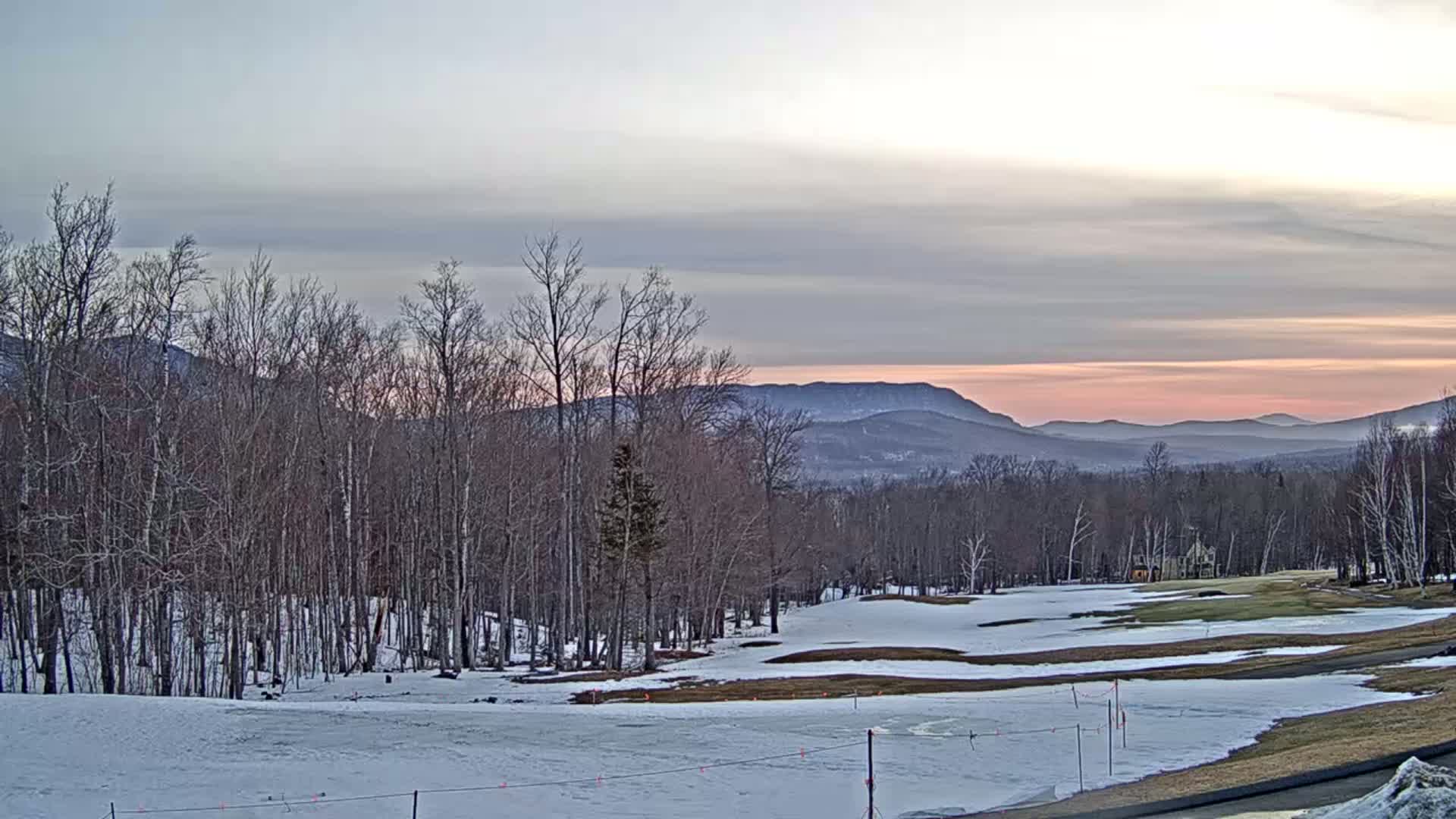 A snow-covered golf course and forest stretch towards a mountain range under a pale pink and grey sunset sky.