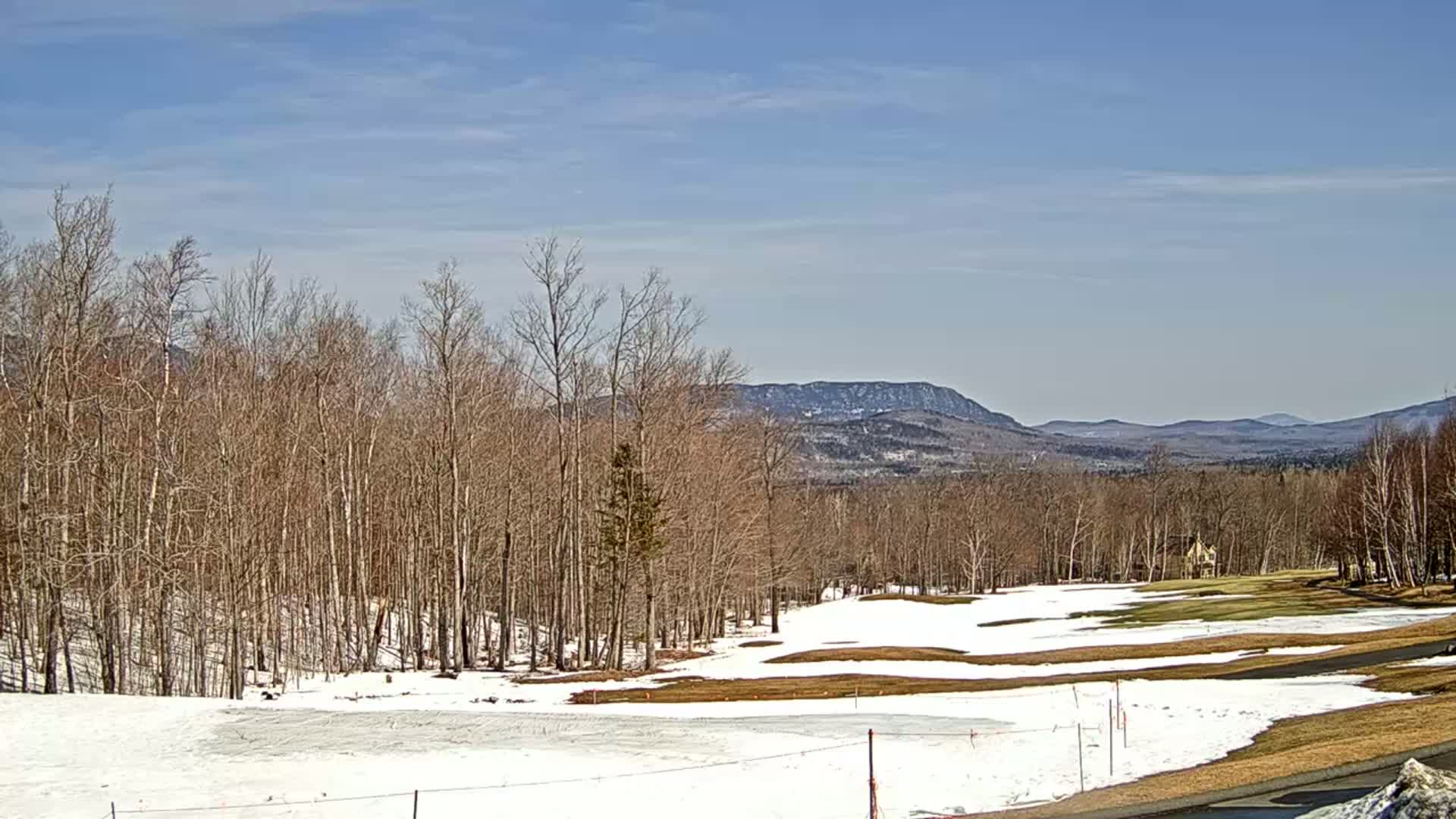 A partially snow-covered golf course sits before a line of bare trees and distant mountains under a mostly sunny sky.
