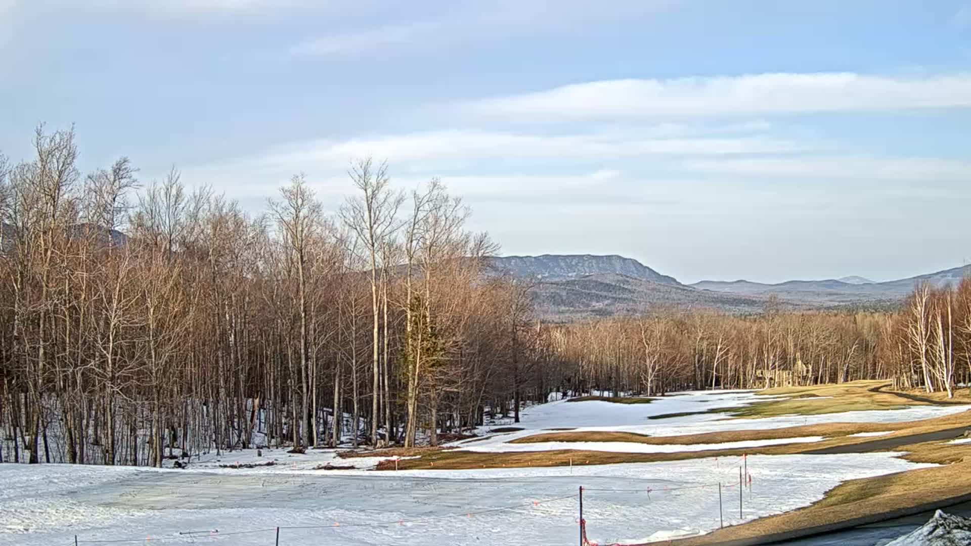 A partly snowy golf course is nestled among bare deciduous trees, with distant mountains under a mostly clear, bright sky.