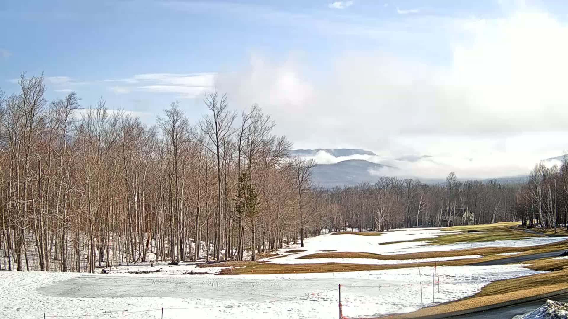 A partly cloudy, winter day reveals a snow-covered golf course nestled amongst bare trees, with distant mountains partially obscured by fog.