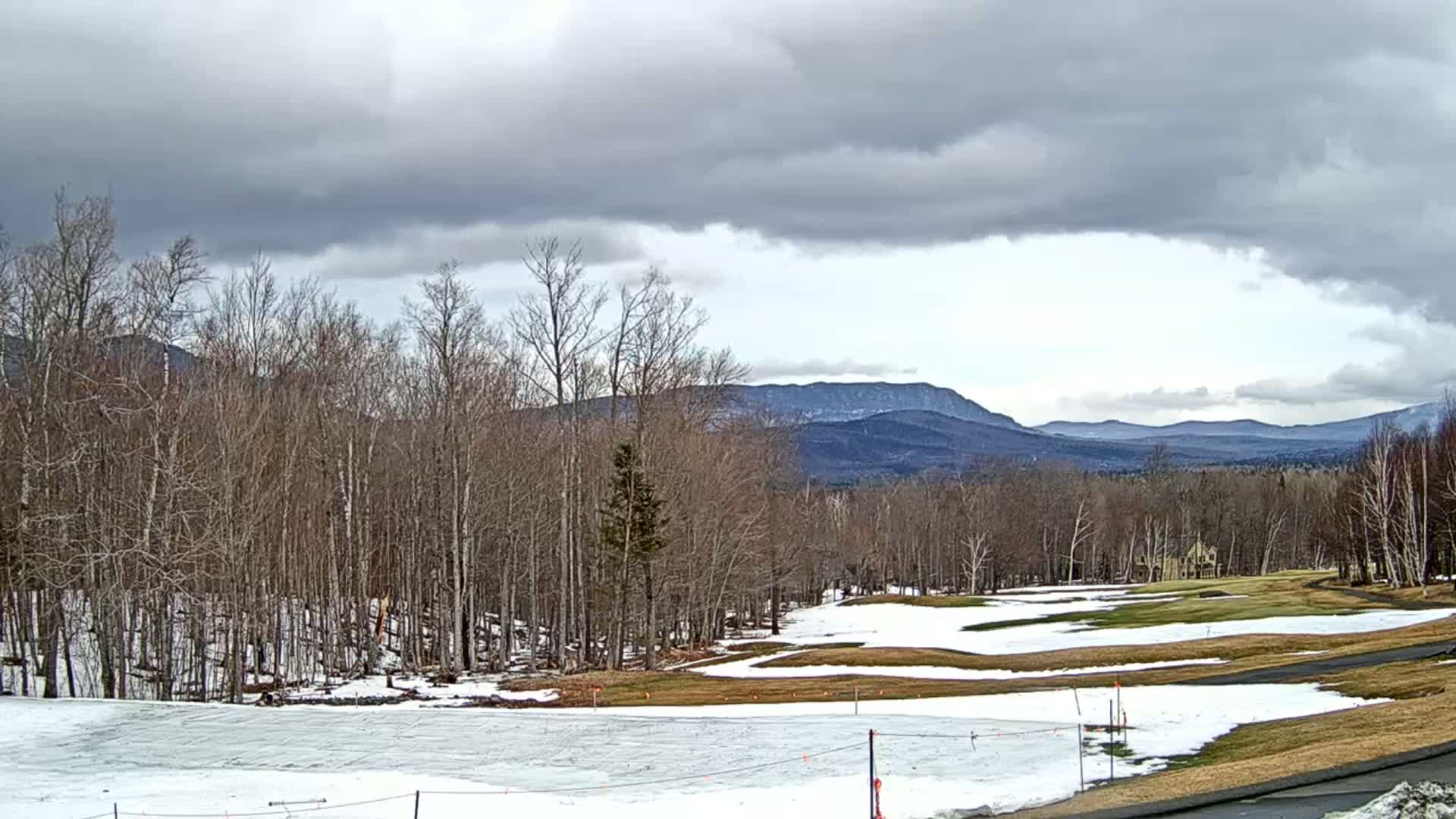 A partially snow-covered golf course sits before a line of bare trees and distant mountains under a cloudy sky.