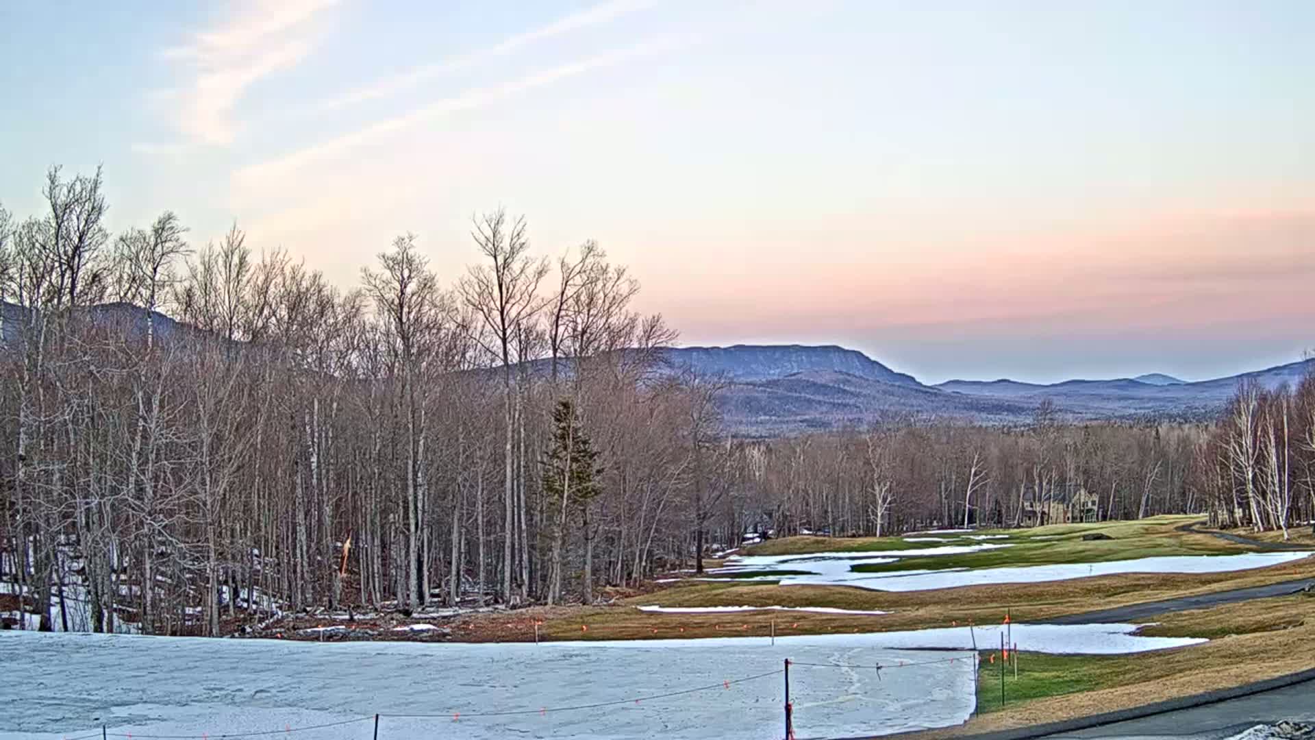 A partly snowy golf course is situated in front of a line of bare deciduous trees and distant mountains under a pale pink and blue sunset sky.