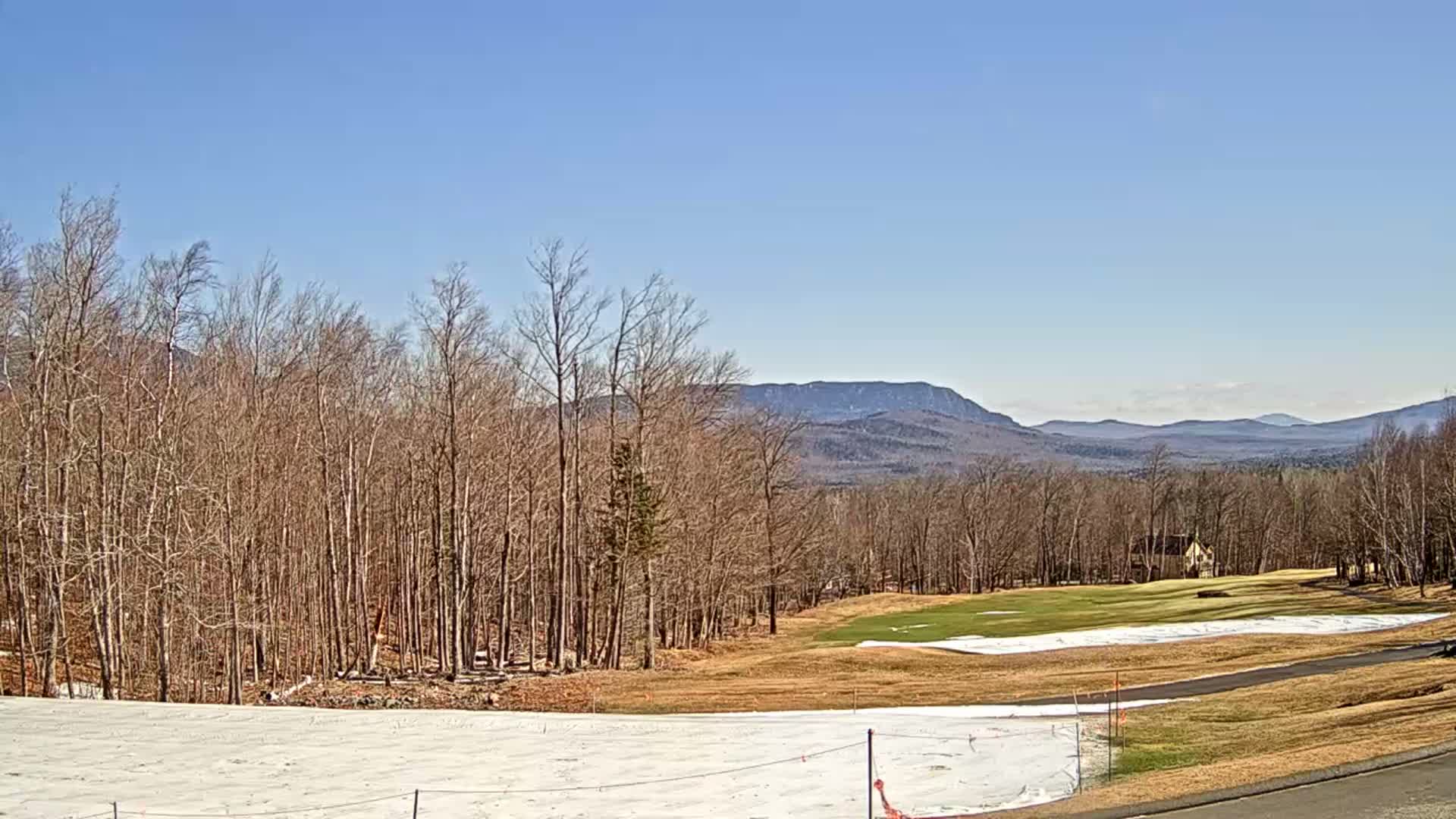 A mostly clear, sunny day reveals a golf course partially covered in snow, bordered by bare deciduous trees and distant mountains.