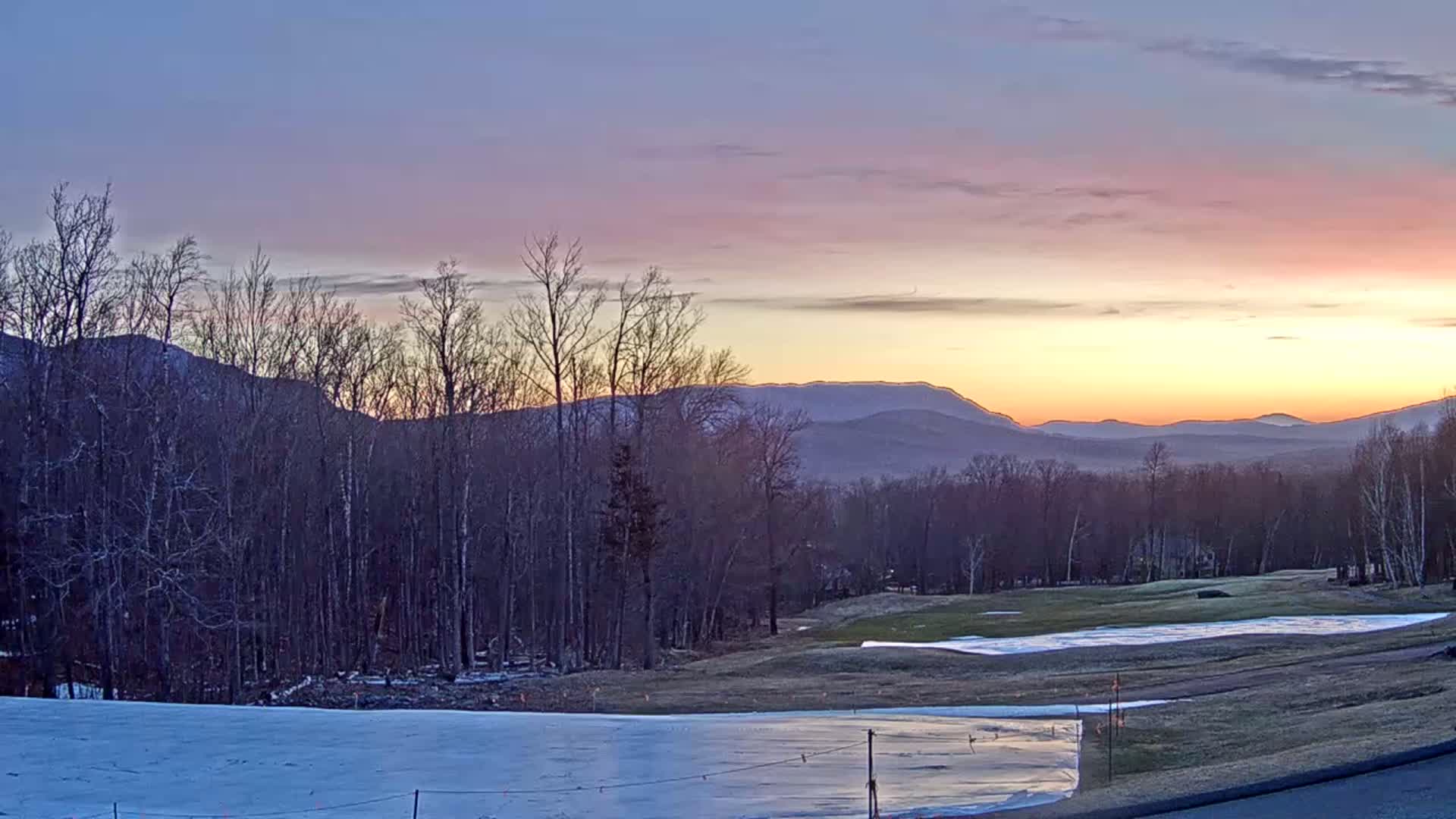 A partly snowy golf course lies in the foreground, with a line of bare trees separating it from distant mountains bathed in the pastel colors of sunrise.