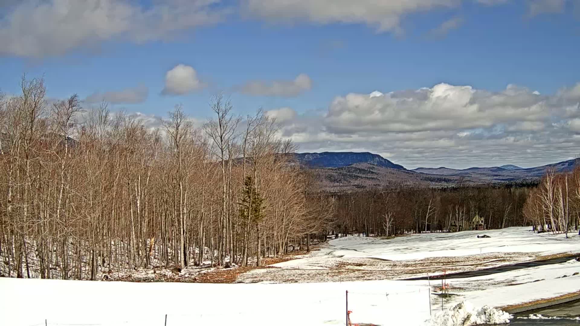 A partly cloudy day reveals a snow-covered landscape with bare, brown trees in the foreground, leading to distant mountains under a mostly blue sky.