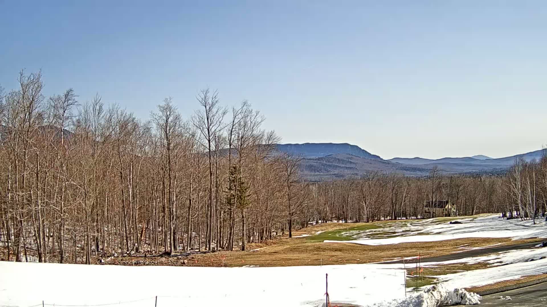 A snow-dusted, barren landscape reveals a golf course nestled amongst leafless trees, with distant mountains under a clear blue sky.