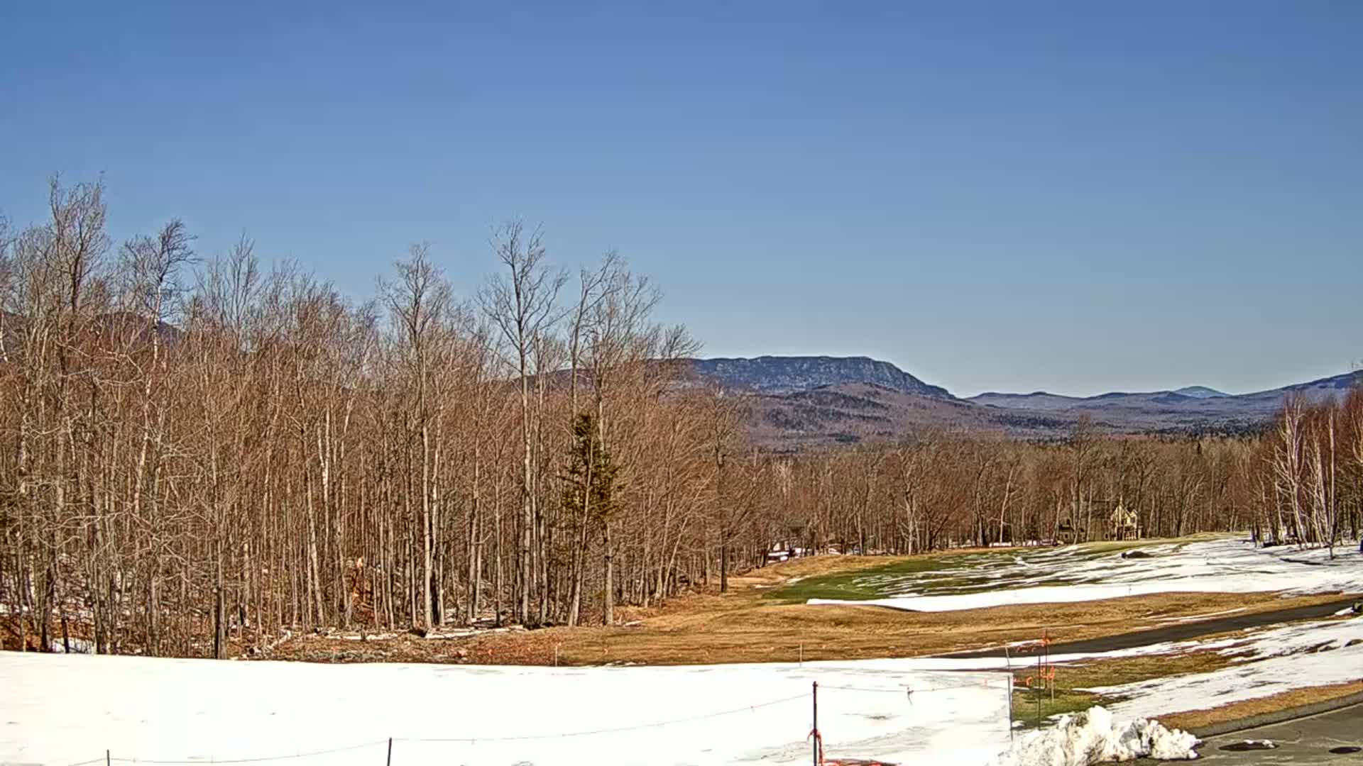 A partly snow-covered landscape with bare trees, a golf course, and distant mountains under a clear blue sky.