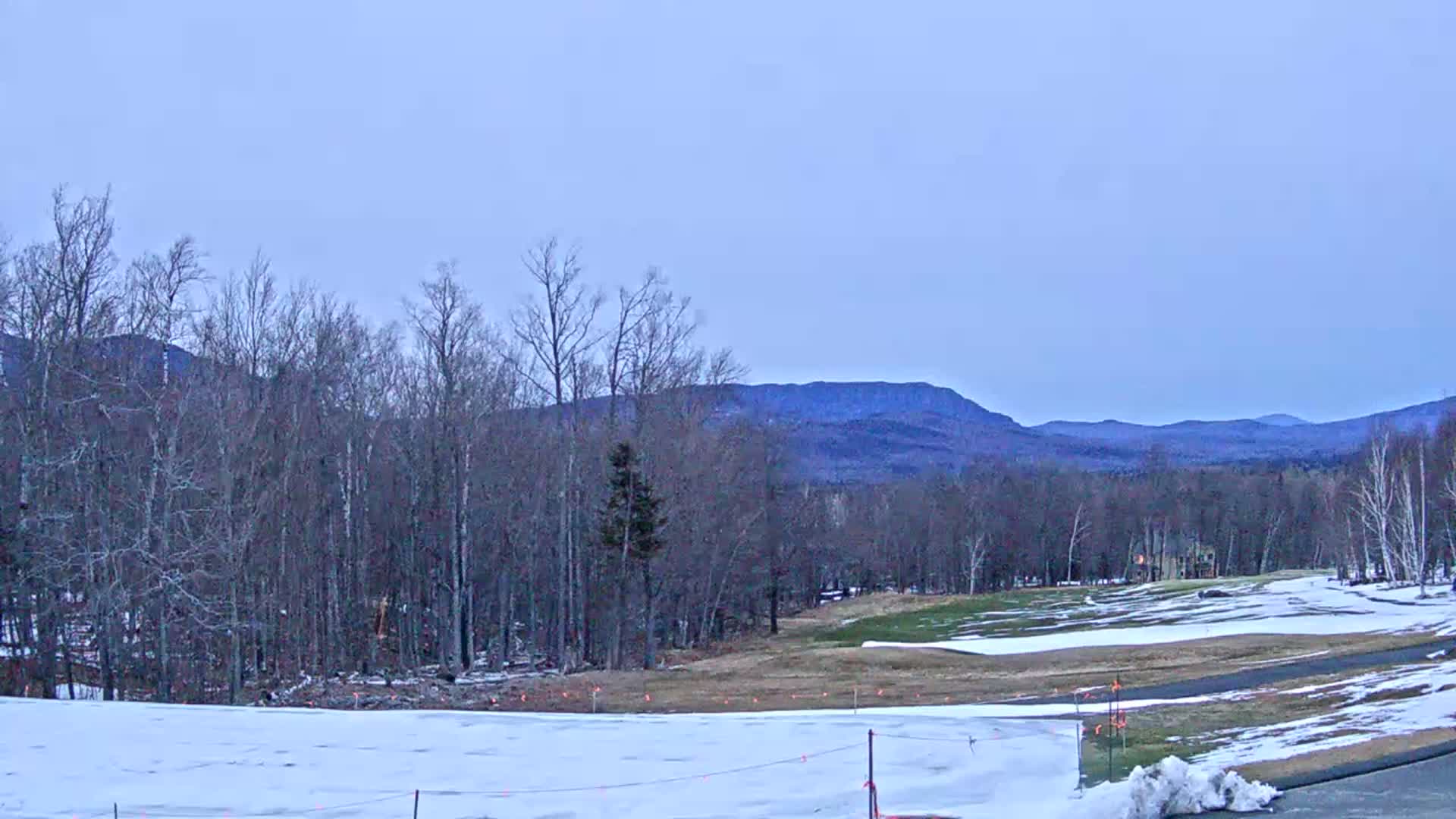 A snow-covered landscape features a partially snow-covered golf course in the foreground, bare trees, and distant mountains under a pale blue, clear sky.