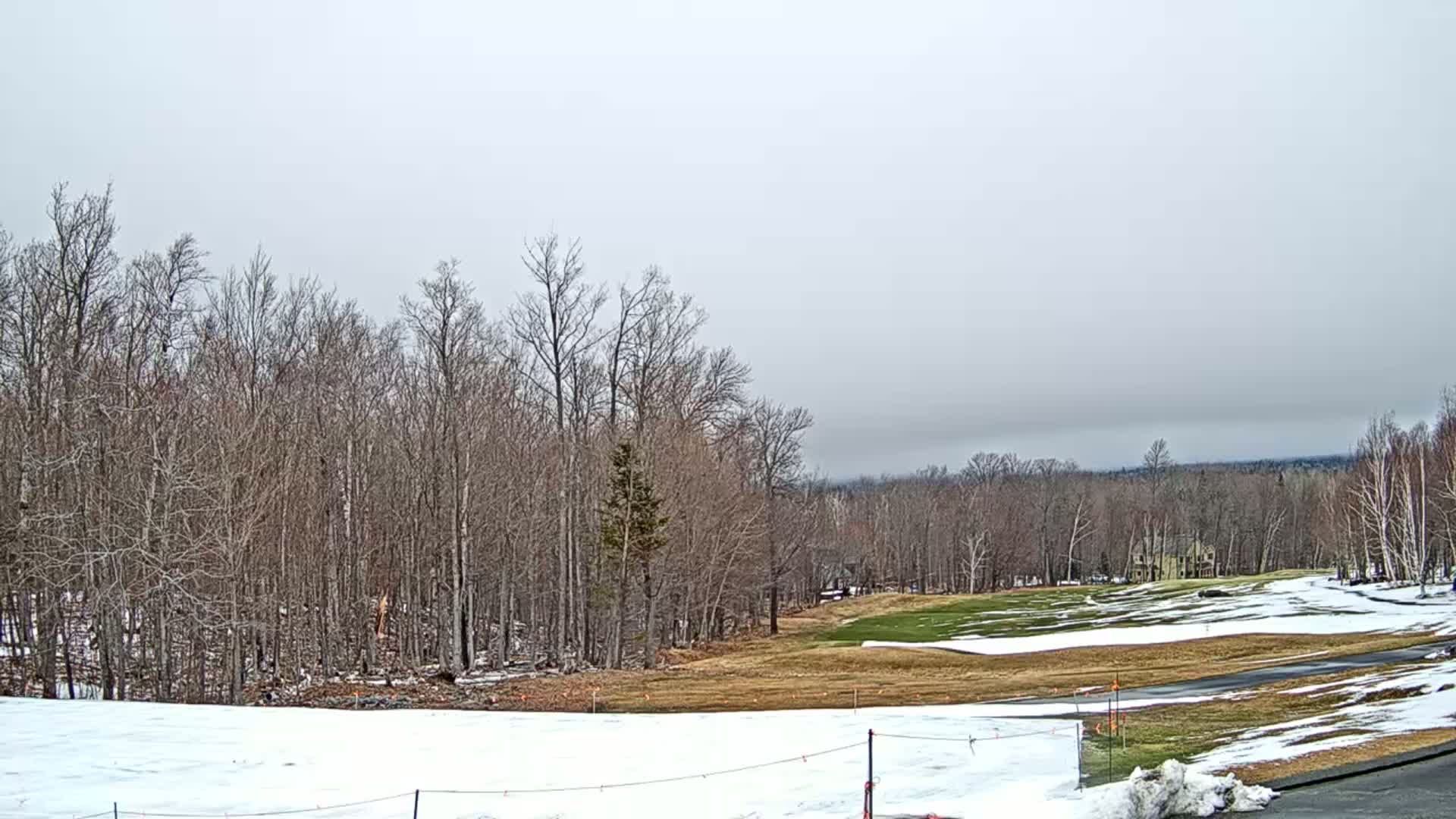 A partially snow-covered golf course is nestled amongst bare, deciduous trees under a cloudy sky.