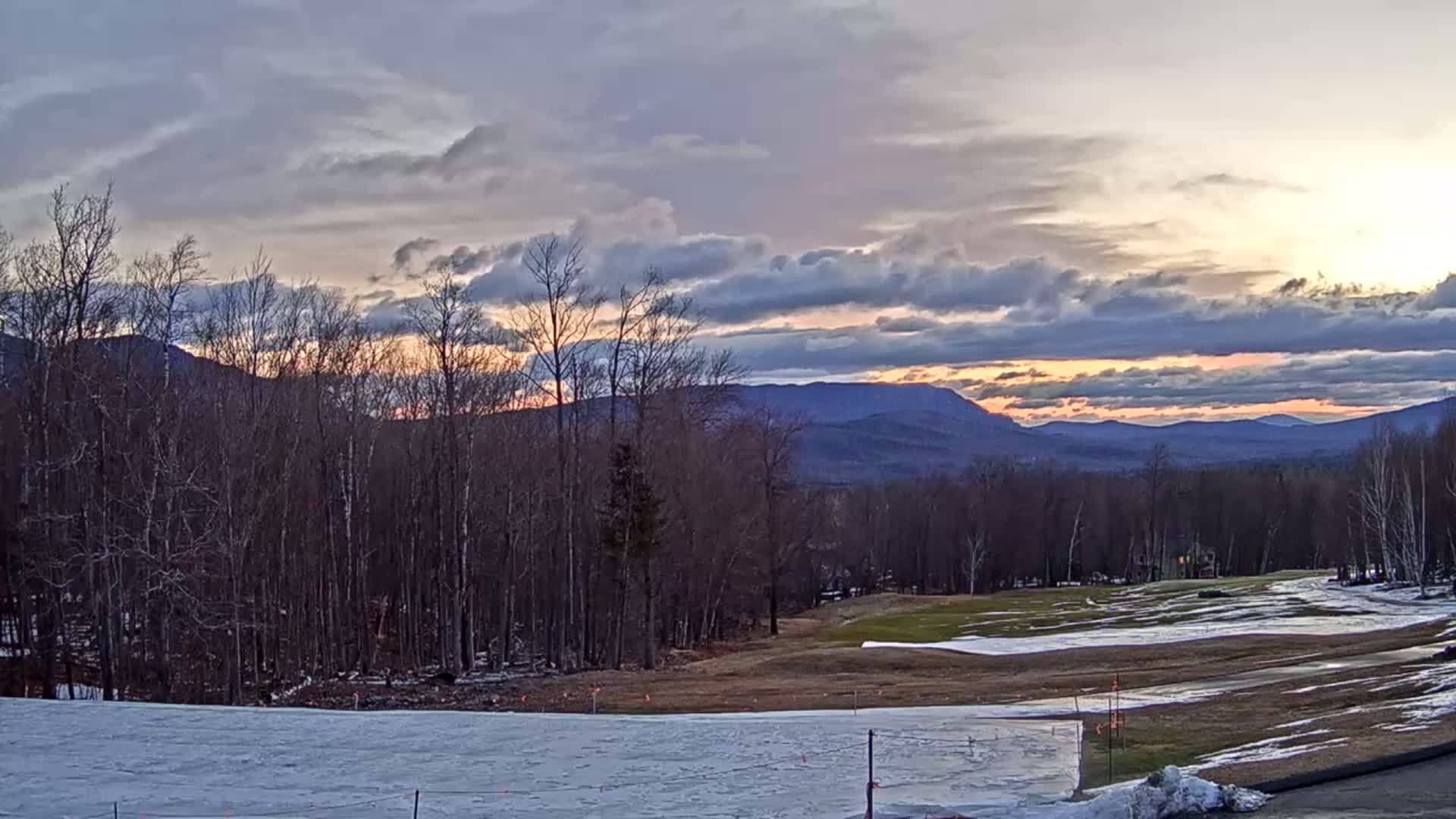 A partly cloudy sunset casts a warm glow over a snow-covered landscape featuring bare trees, a partially melted golf course, and distant mountains.
