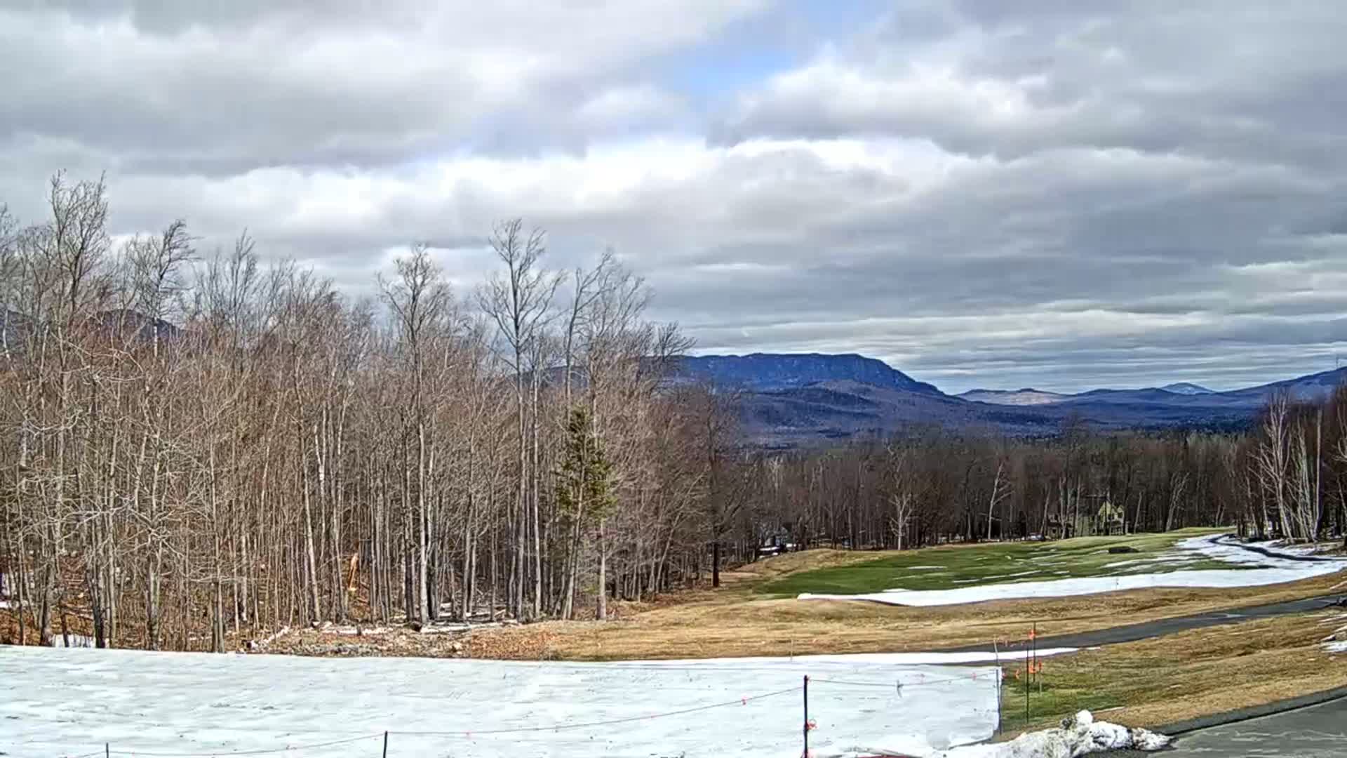 A partly snow-covered golf course and bare deciduous trees stretch towards distant mountains under a cloudy sky.