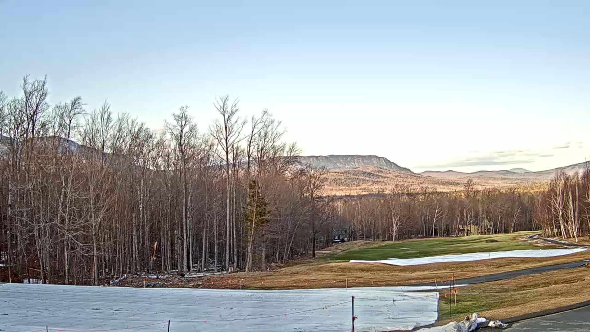 A partially snow-covered golf course and bare deciduous trees sit before distant, snow-capped mountains under a clear, light blue sky.