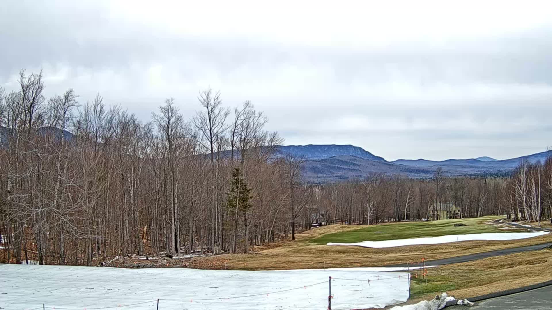 A partially snow-covered golf course lies in the foreground, with bare deciduous trees and distant mountains under a cloudy sky.