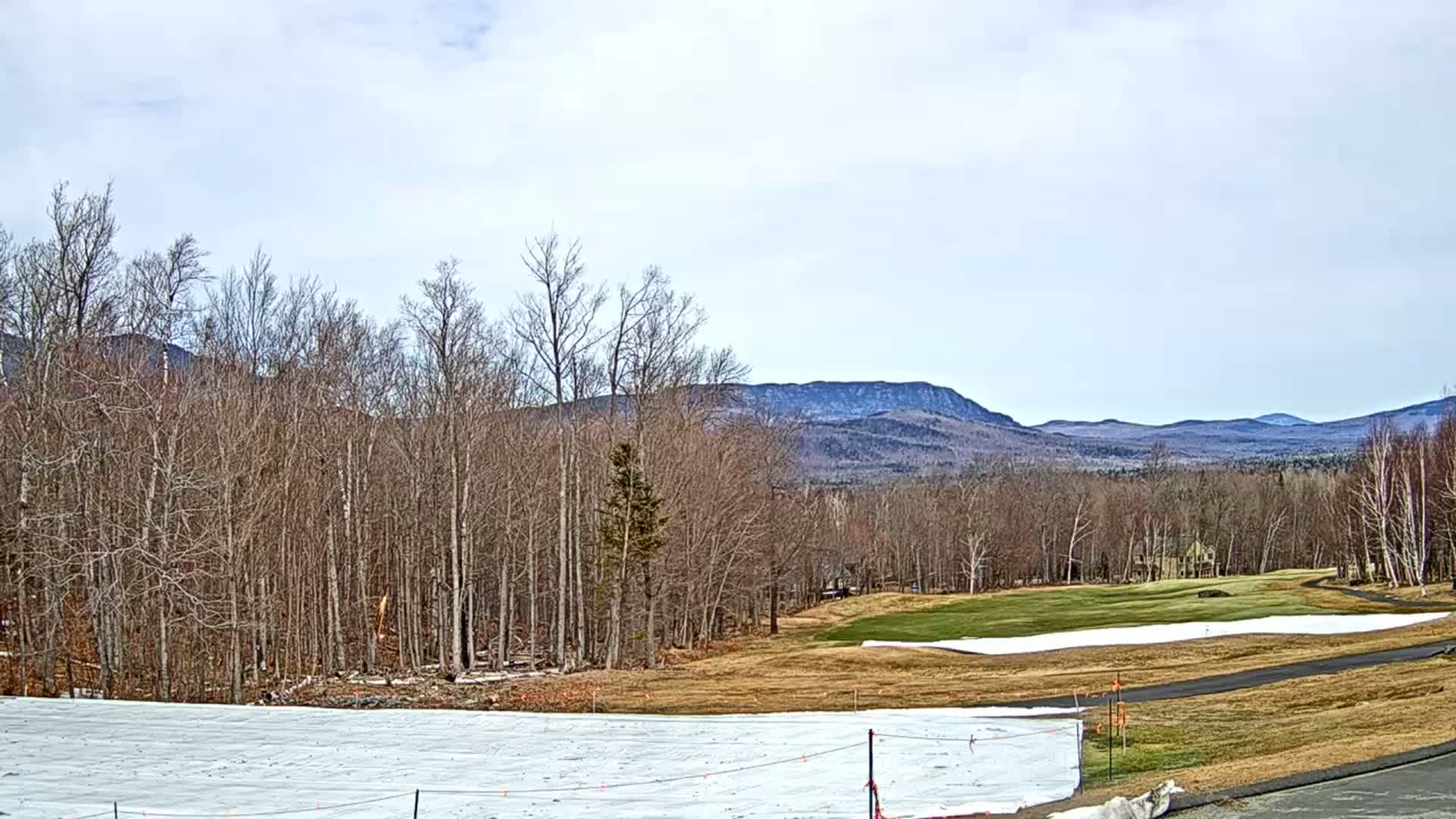 A mostly cloudy day reveals a view of a golf course partially covered in snow, bare deciduous trees, and distant mountains.