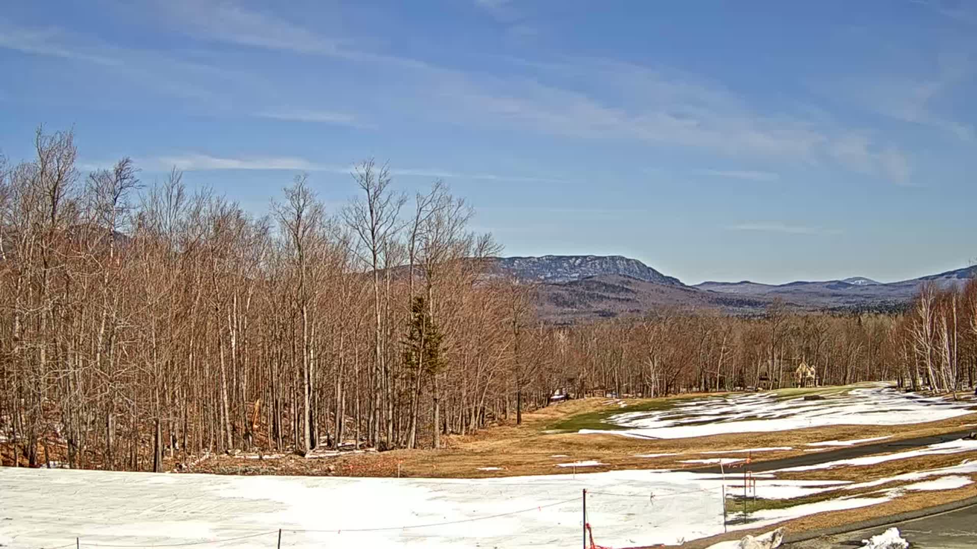 A mostly clear sky overlooks a snow-dusted golf course bordered by a forest of leafless trees and distant mountains.