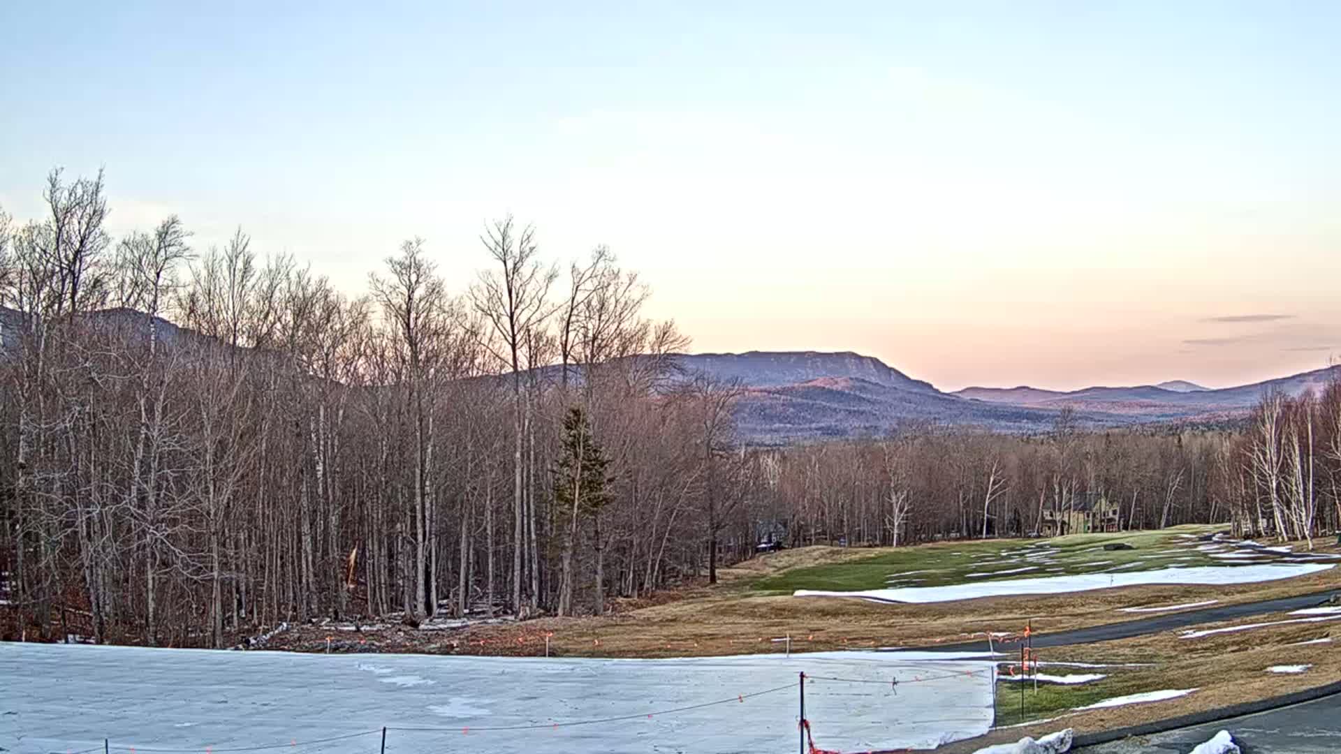 A partially snow-covered golf course and bare deciduous trees sit before a range of mountains under a pale pink and blue twilight sky.