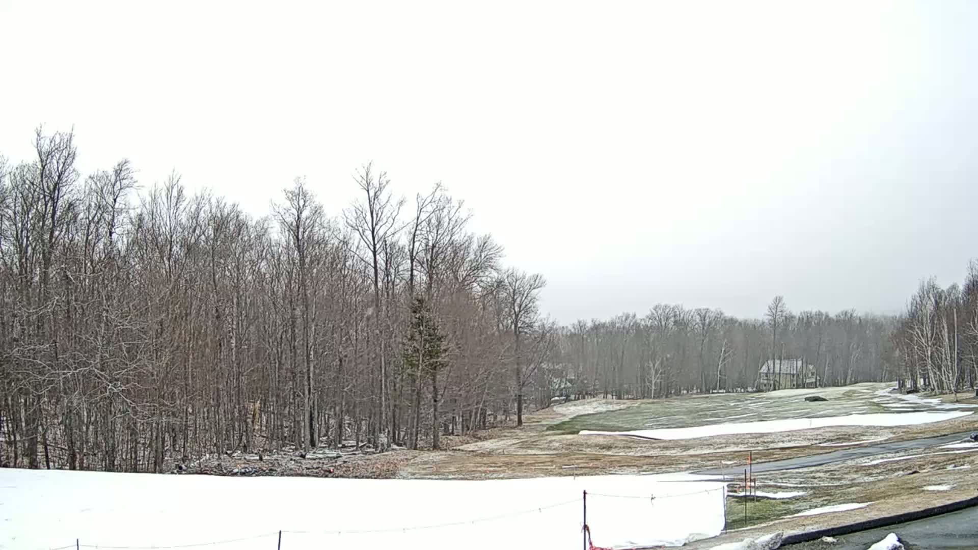 A partly snow-covered golf course is nestled amongst leafless trees under an overcast sky.