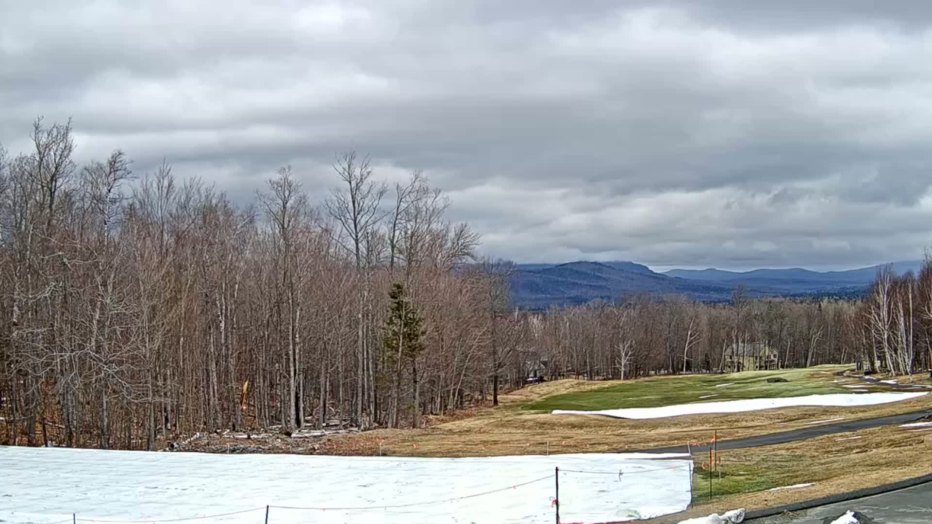 A partly snow-covered golf course sits before a line of leafless trees, with distant blue mountains under a cloudy sky.