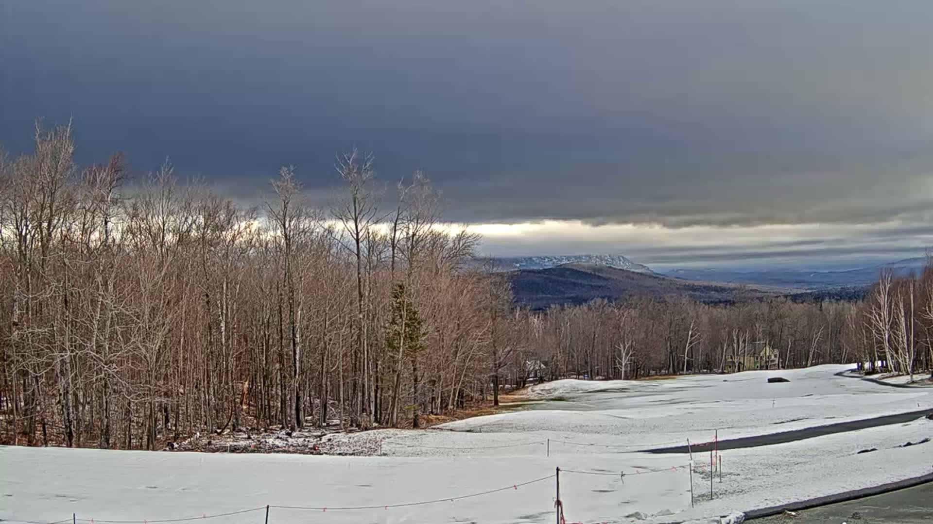 A snow-covered golf course and bare deciduous forest are visible beneath a dark, cloudy sky, with distant mountains in the background.