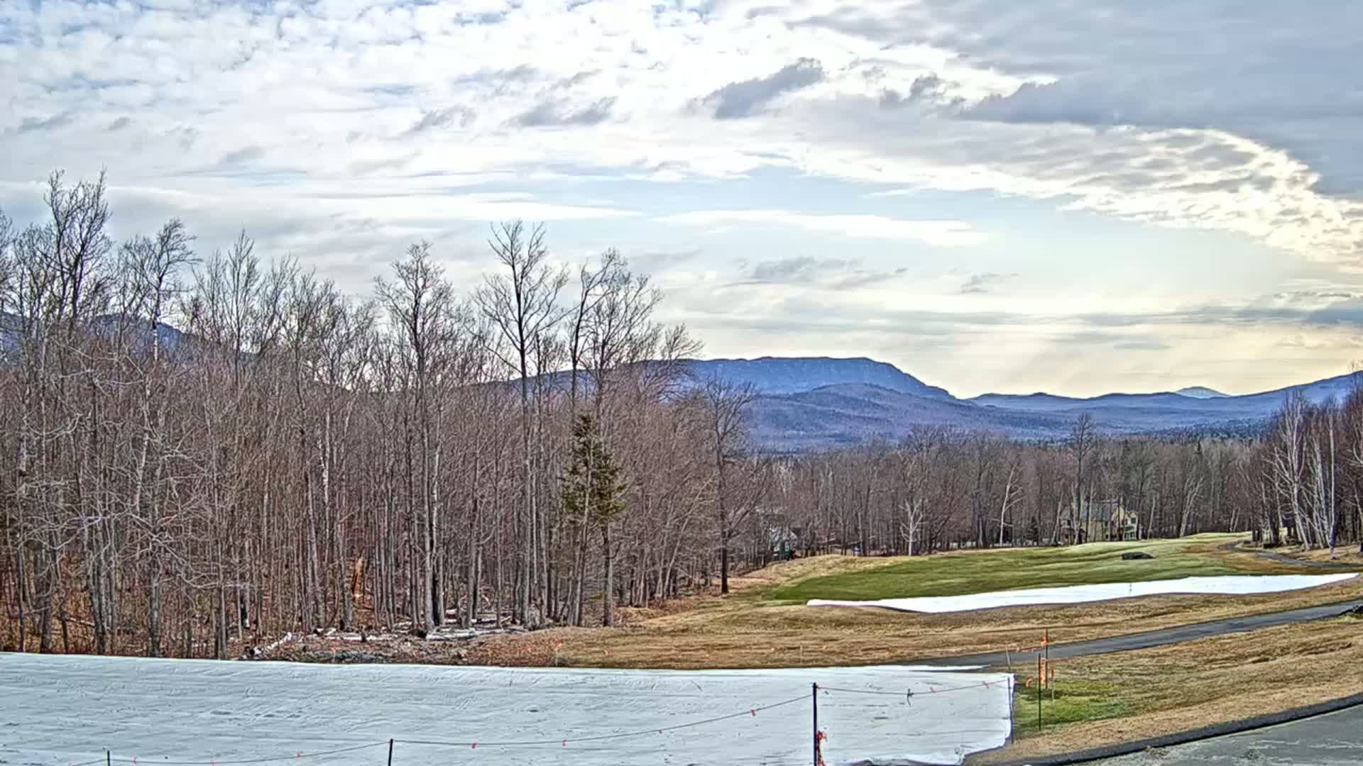 A mostly cloudy sky overlooks a snow-covered field, a partially snow-covered golf course, and bare deciduous trees leading to distant, hazy mountains.