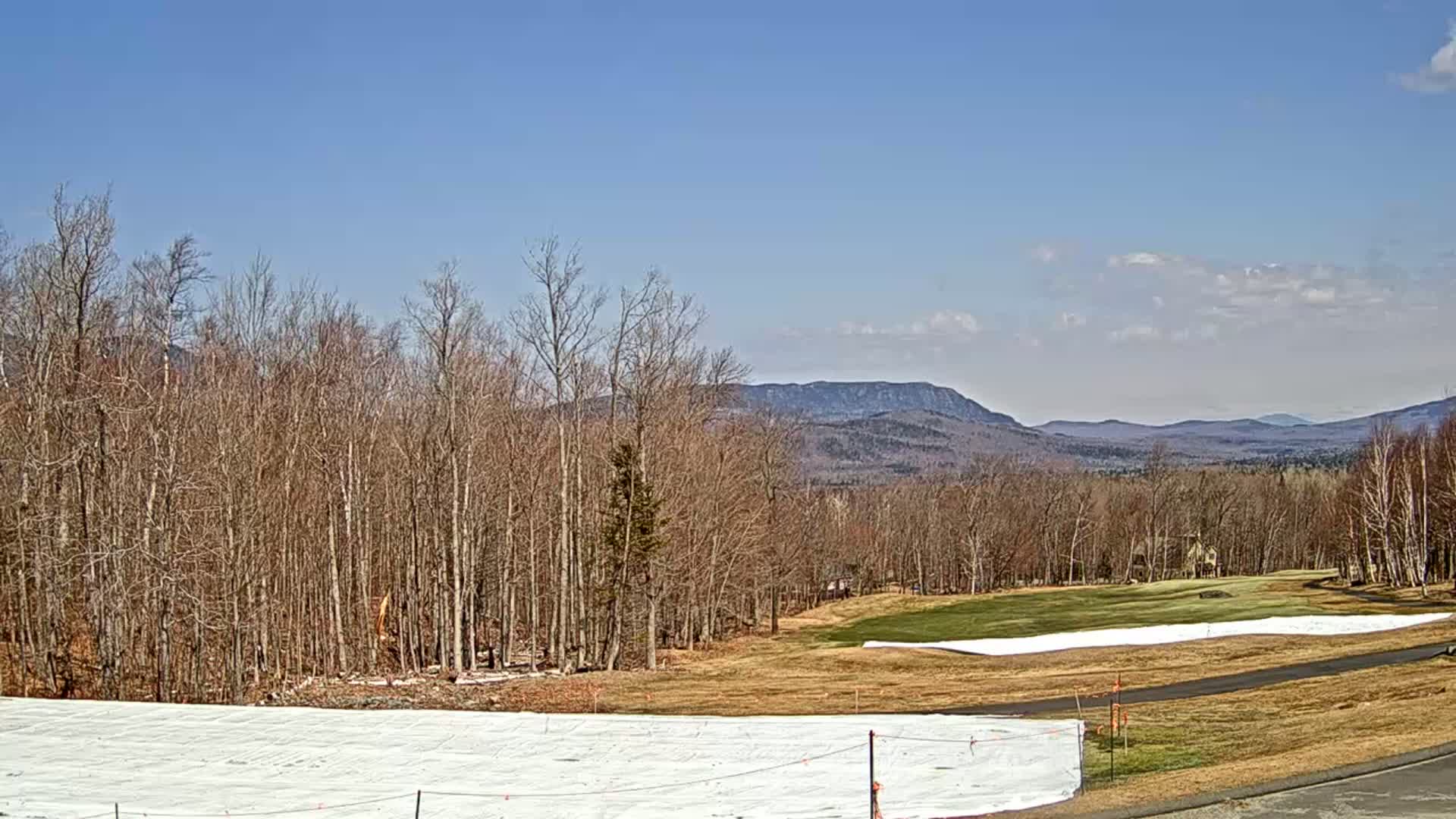 A partially snow-covered golf course and barren trees stretch toward distant mountains under a clear blue sky with a few clouds.
