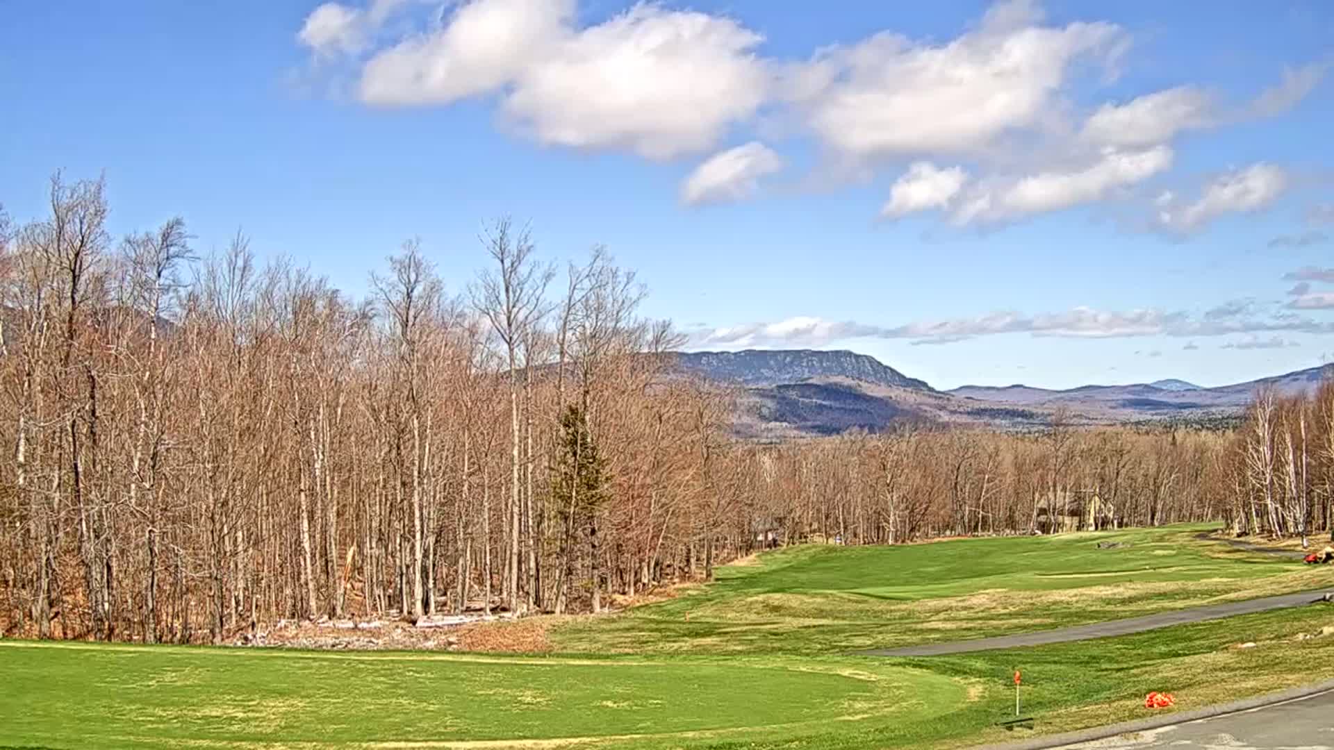 A mostly sunny day reveals a golf course nestled among bare deciduous trees and mountains in the distance.