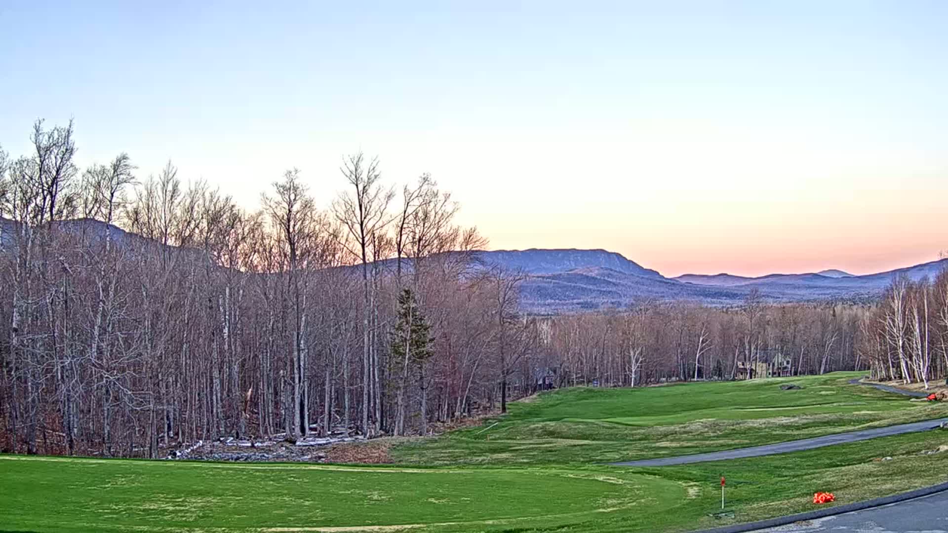 A golf course with green grass and a paved path stretches towards a line of bare trees and distant mountains under a pale pink and blue sunset sky.