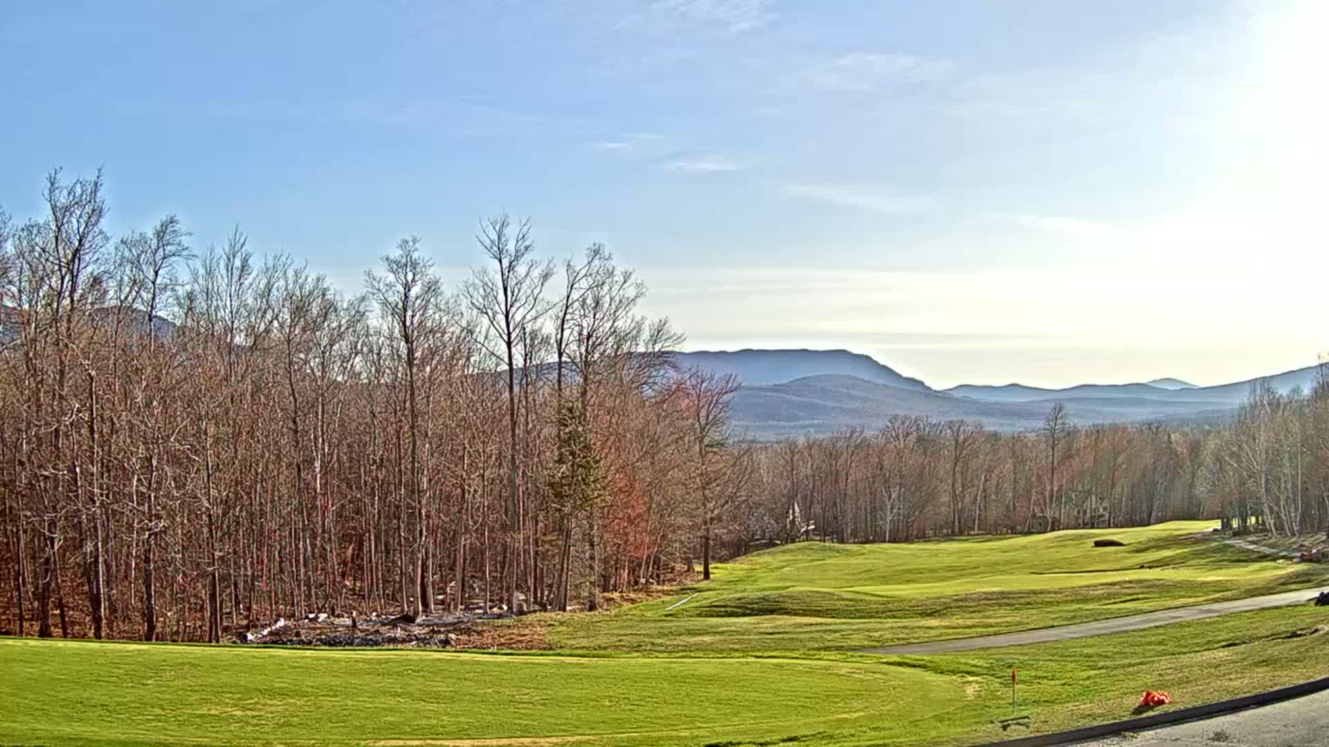 A golf course with a mostly bare deciduous forest in the foreground and distant mountains under a clear, bright sky.