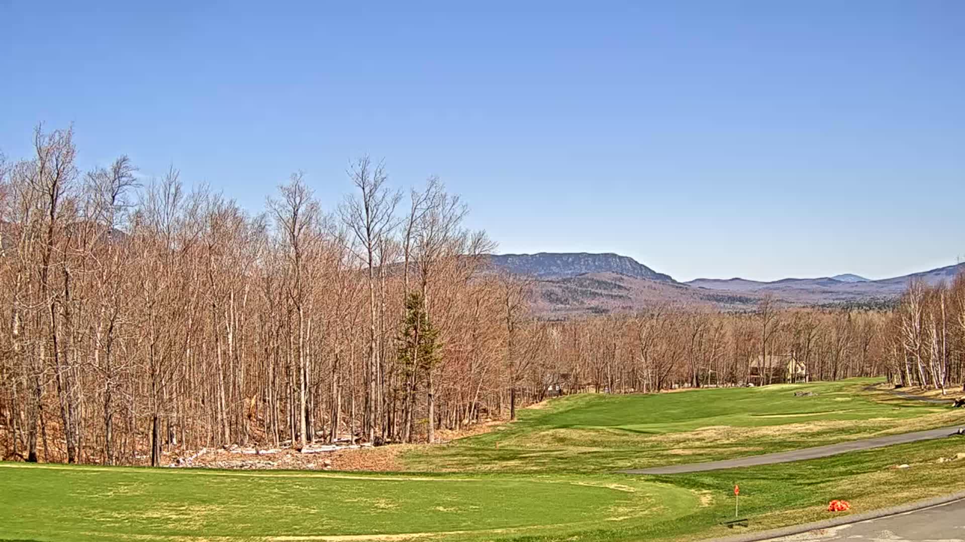 A sunny day reveals a golf course nestled among bare trees, with distant mountains under a clear blue sky.