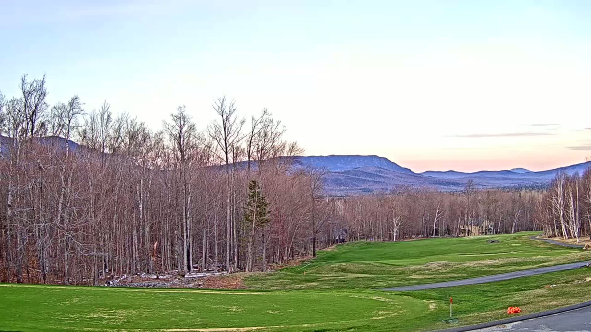 A golf course with a green fairway and a road curves around it,  bare trees in the foreground, and distant mountains under a pale pink and blue sky.