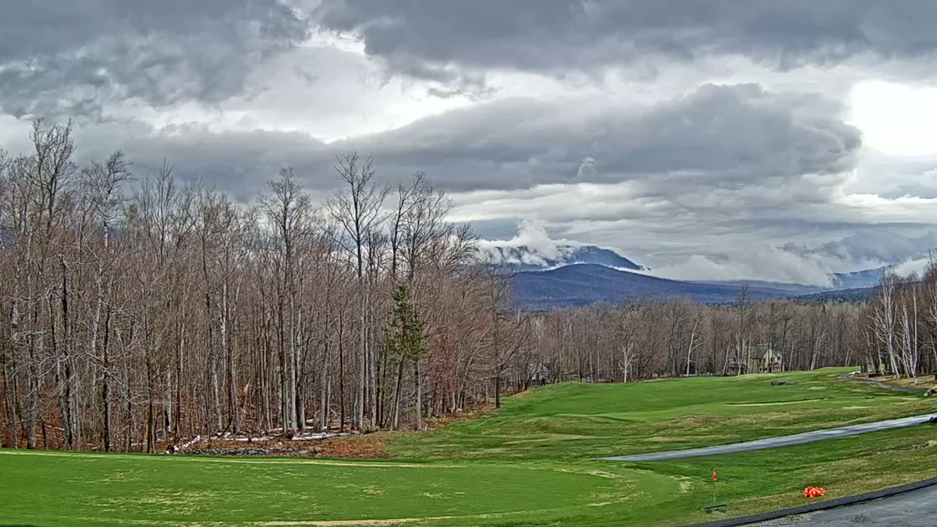 A golf course with a line of bare trees sits in the foreground, overlooking a range of mountains partially obscured by clouds under an overcast sky.