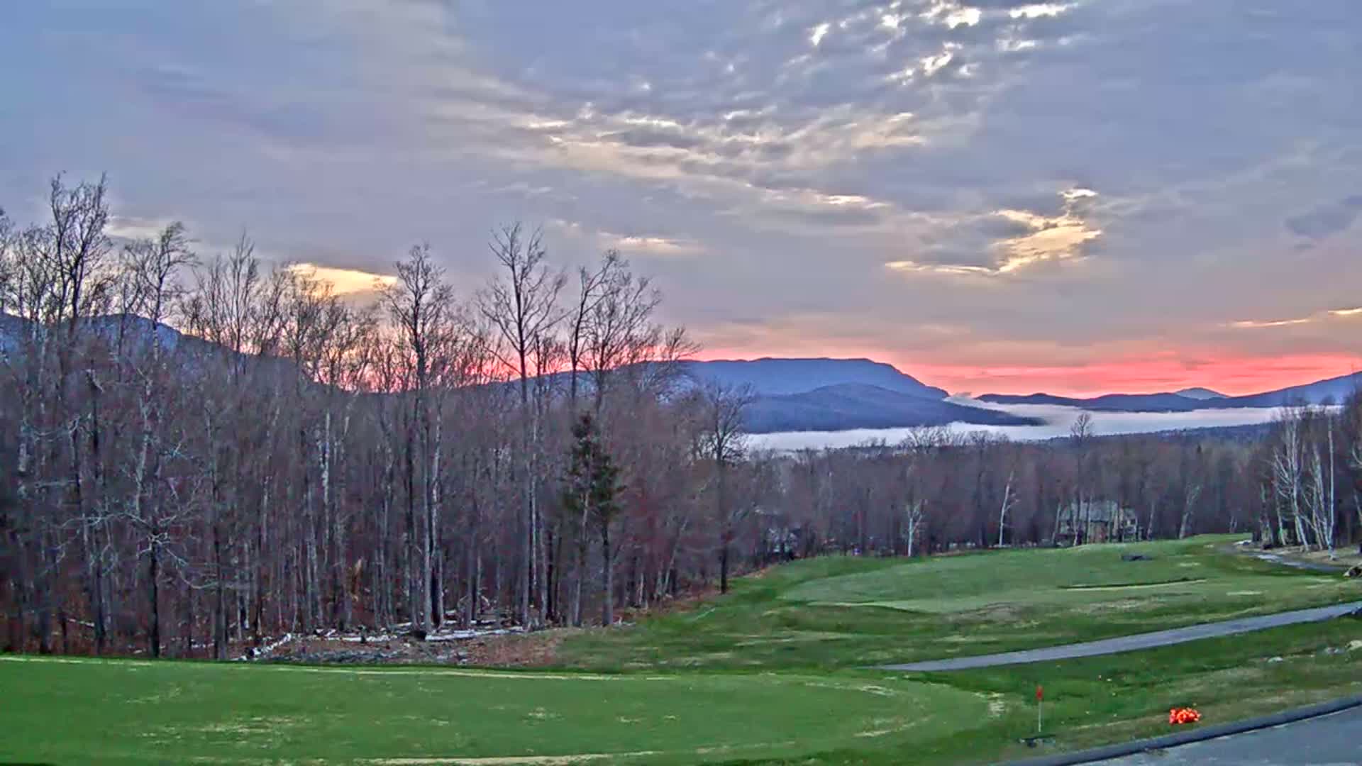 A golf course overlooks a valley filled with fog at sunrise, with mountains in the background under a partly cloudy sky.