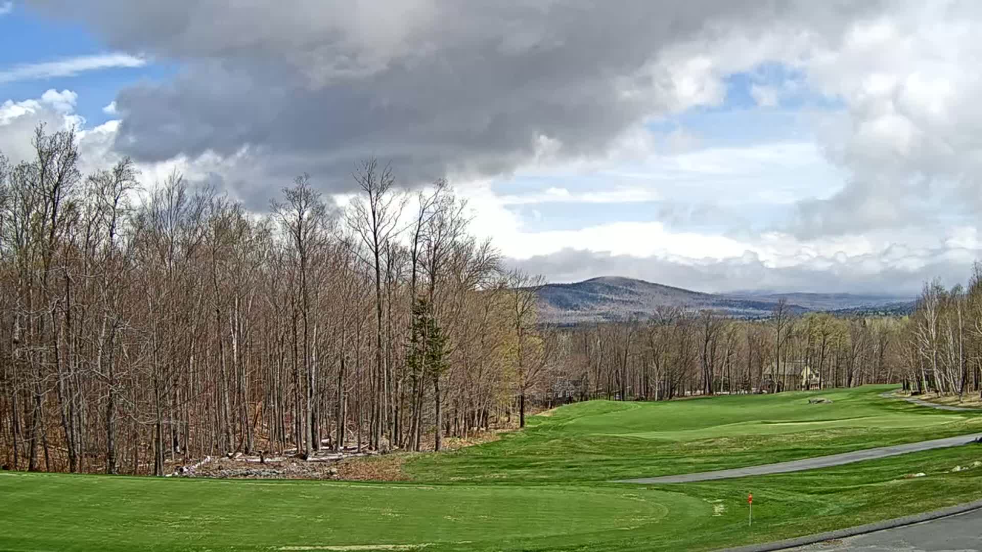 A mostly cloudy day reveals a golf course nestled among bare trees, overlooking distant mountains.