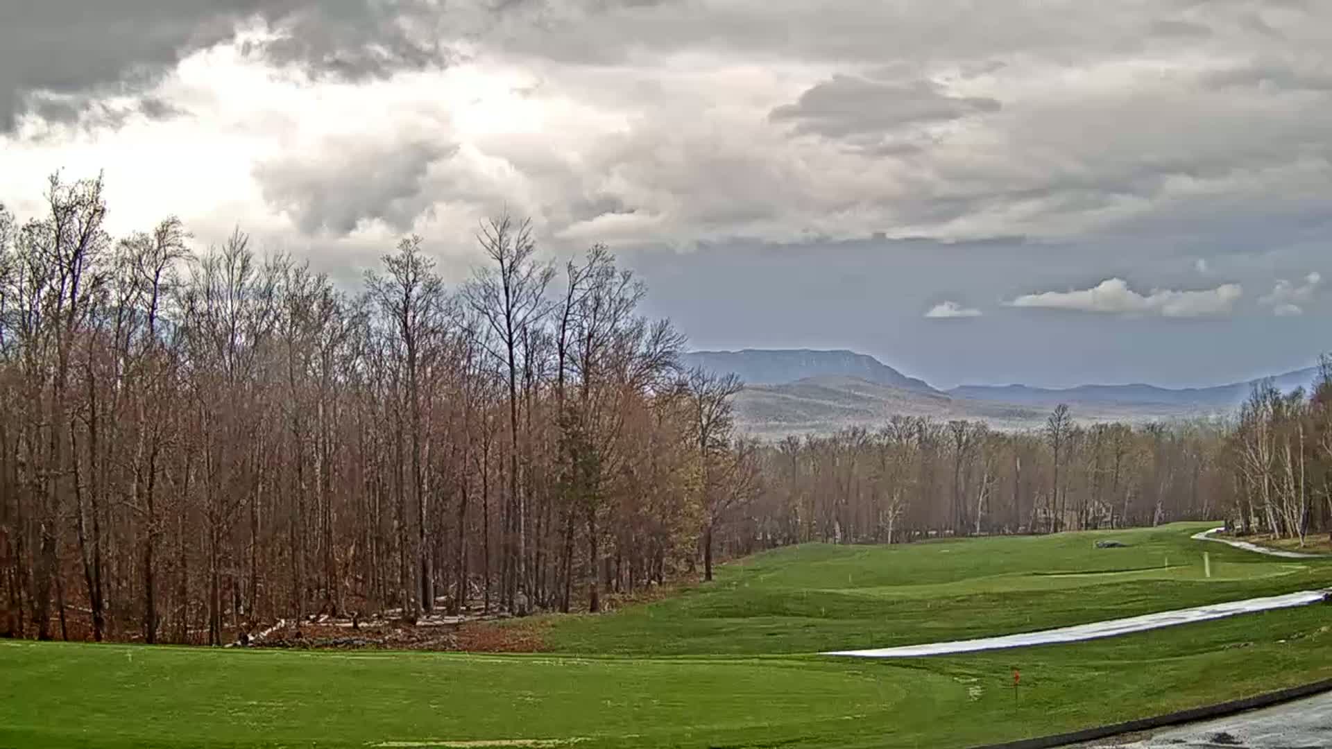 A mostly cloudy day reveals a golf course nestled in a forest with distant mountains visible in the background.