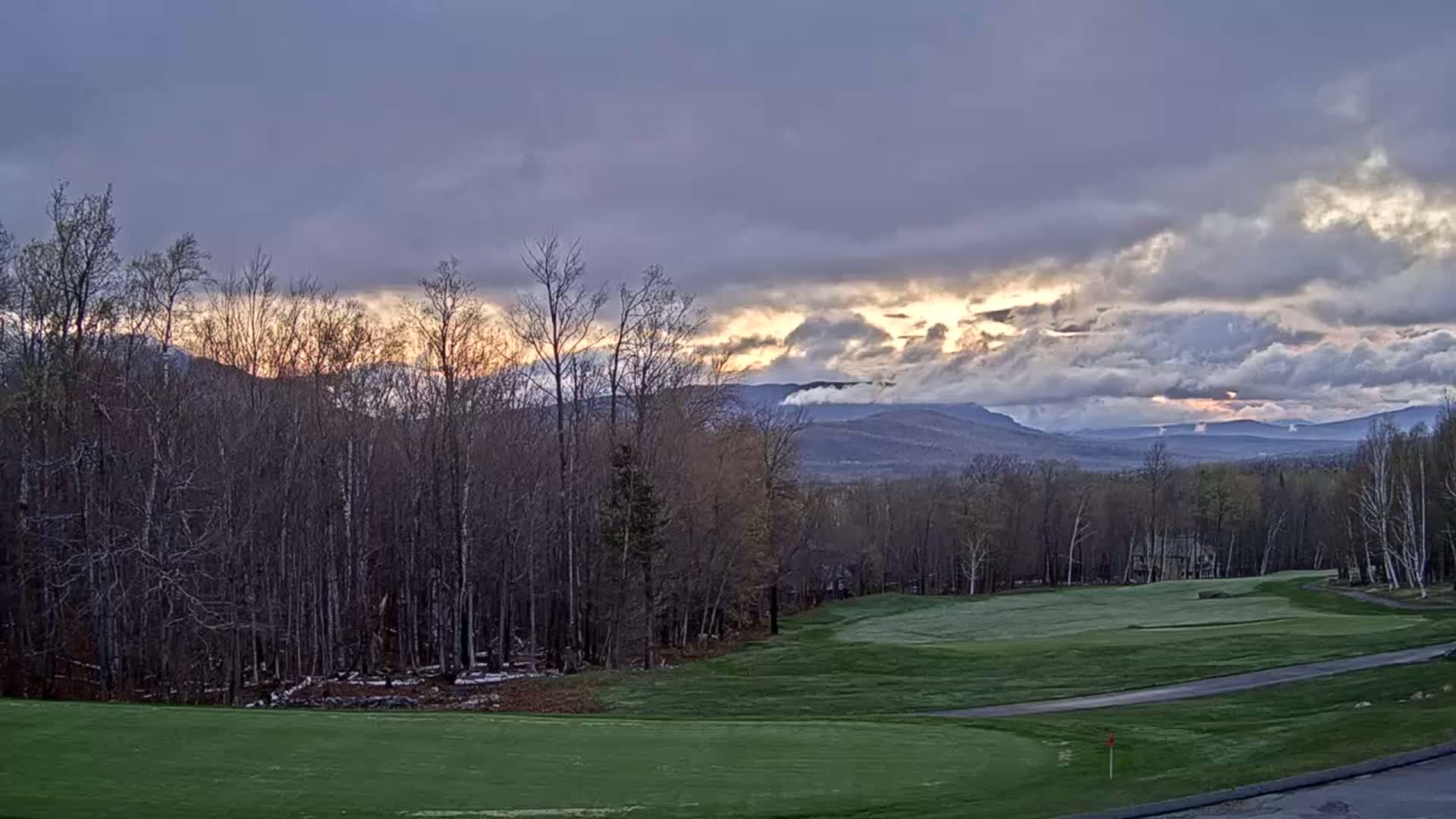A golf course lies before a line of bare trees, with distant mountains visible under a partly cloudy sky at sunrise.